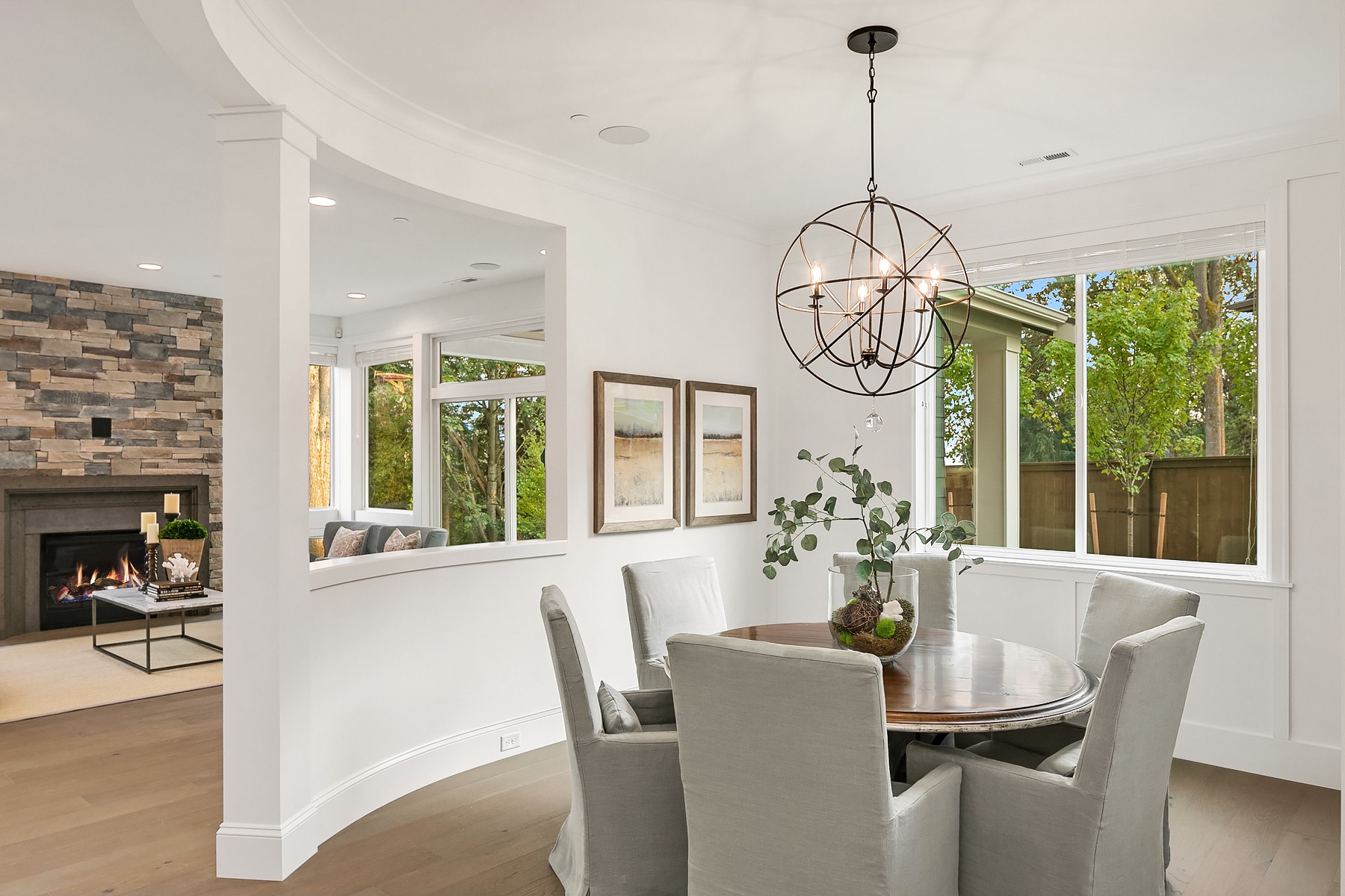 A modern dining room features a round wooden table surrounded by five upholstered chairs. The room is lit by an intricate pendant light fixture. Large windows with views of greenery bring in ample natural light, and a partial wall separates the dining area from the cozy living room.