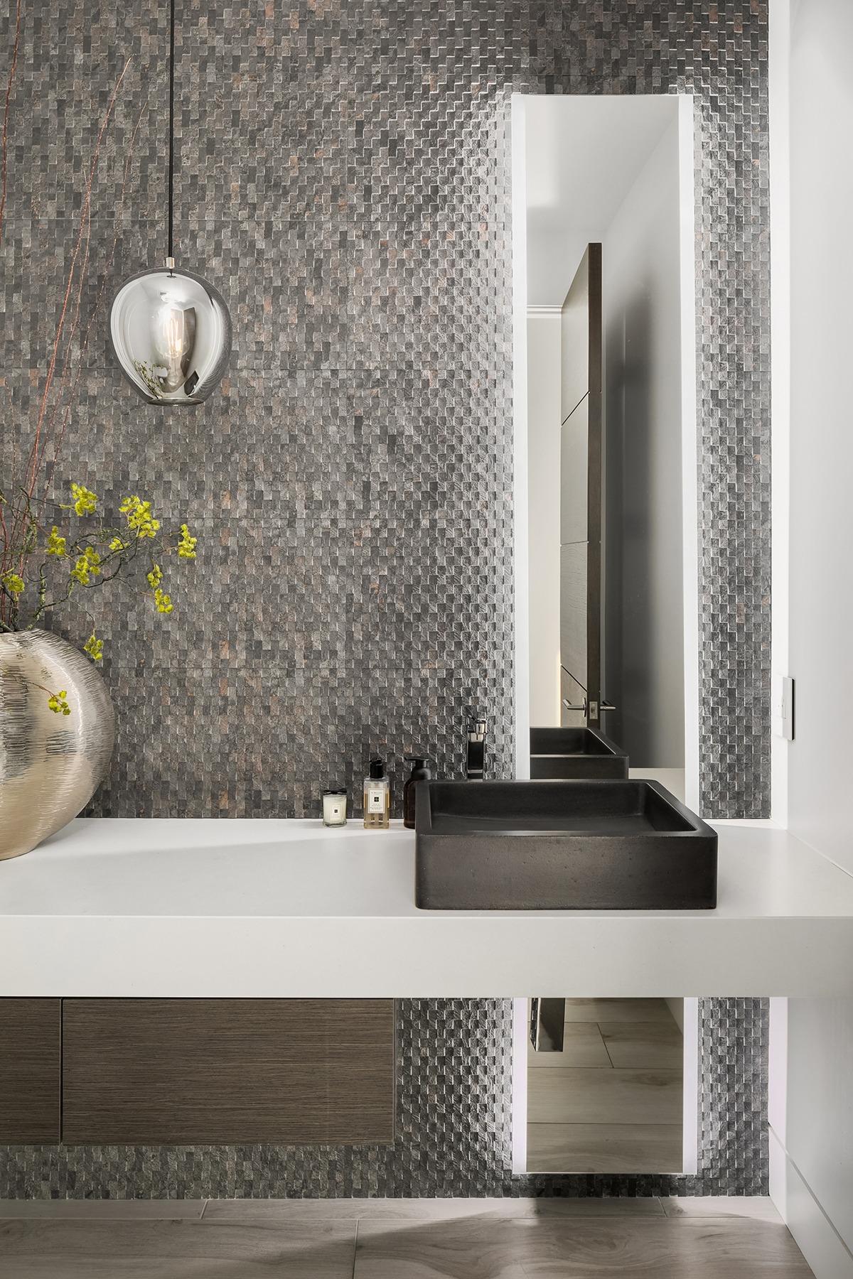 Modern bathroom with textured gray tile wall, sleek white countertop, and black rectangular sink. A spherical pendant light hangs above, and yellow flowers in a large vase decorate the counter. Doorway with open door visible in the background.