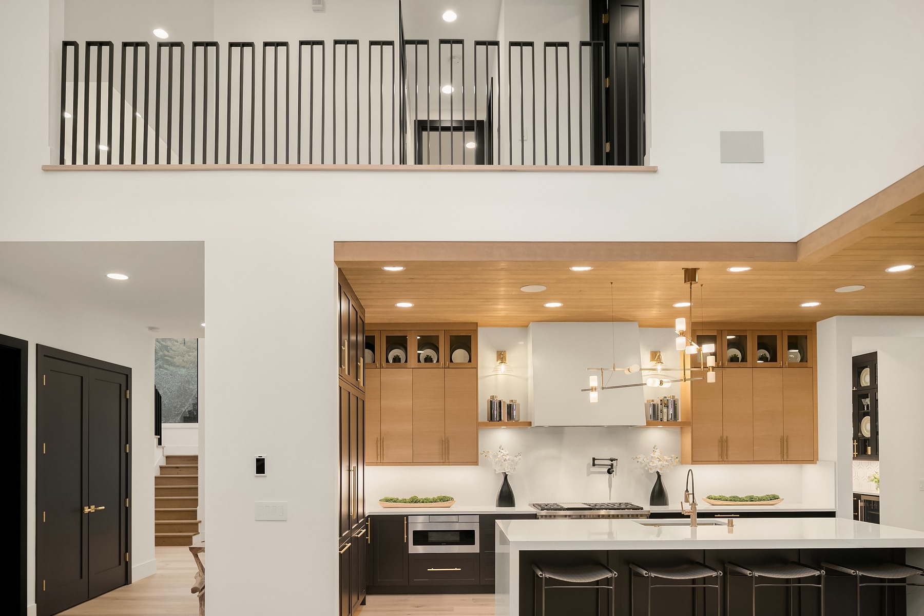 Modern kitchen with a combination of light and dark cabinetry, sleek countertops, and stylish pendant lighting. The space features an island with barstools, and a unique upper walkway with black railings. A staircase is visible to the left. The ceiling is high.