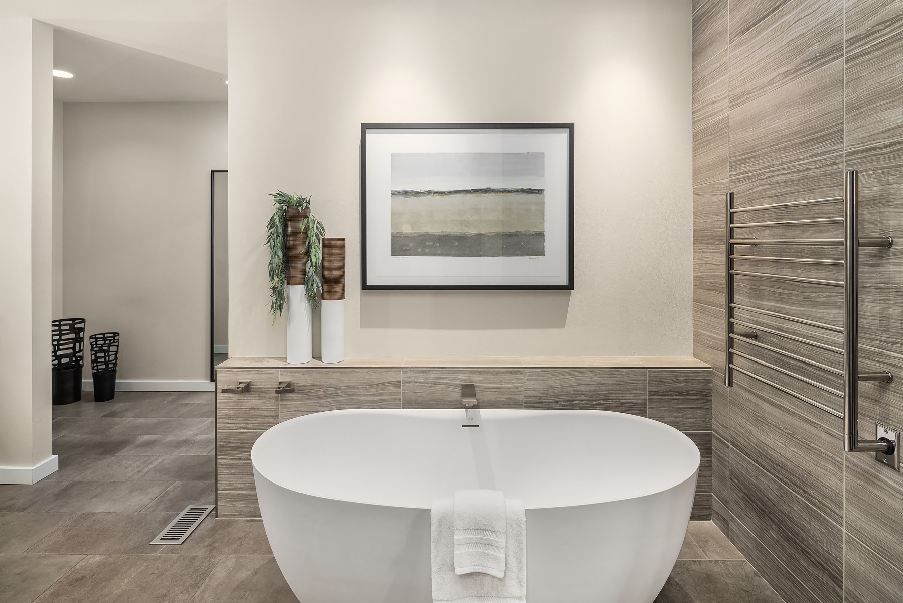 A modern bathroom featuring a freestanding white oval bathtub with a towel draped over its side. Behind the tub, a wall displays a framed landscape artwork and two tall vases with greenery. The bathroom has neutral-toned tiles and floor, with a sleek, minimalist design.