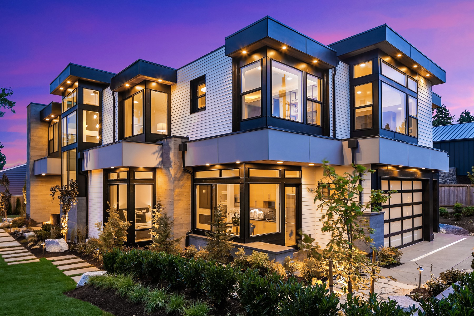 A modern two-story house with large windows, glowing warmly from interior lights, set against a purple twilight sky. The exterior features a mix of white siding and dark accents. A landscaped garden and pathway lead to the entrance.