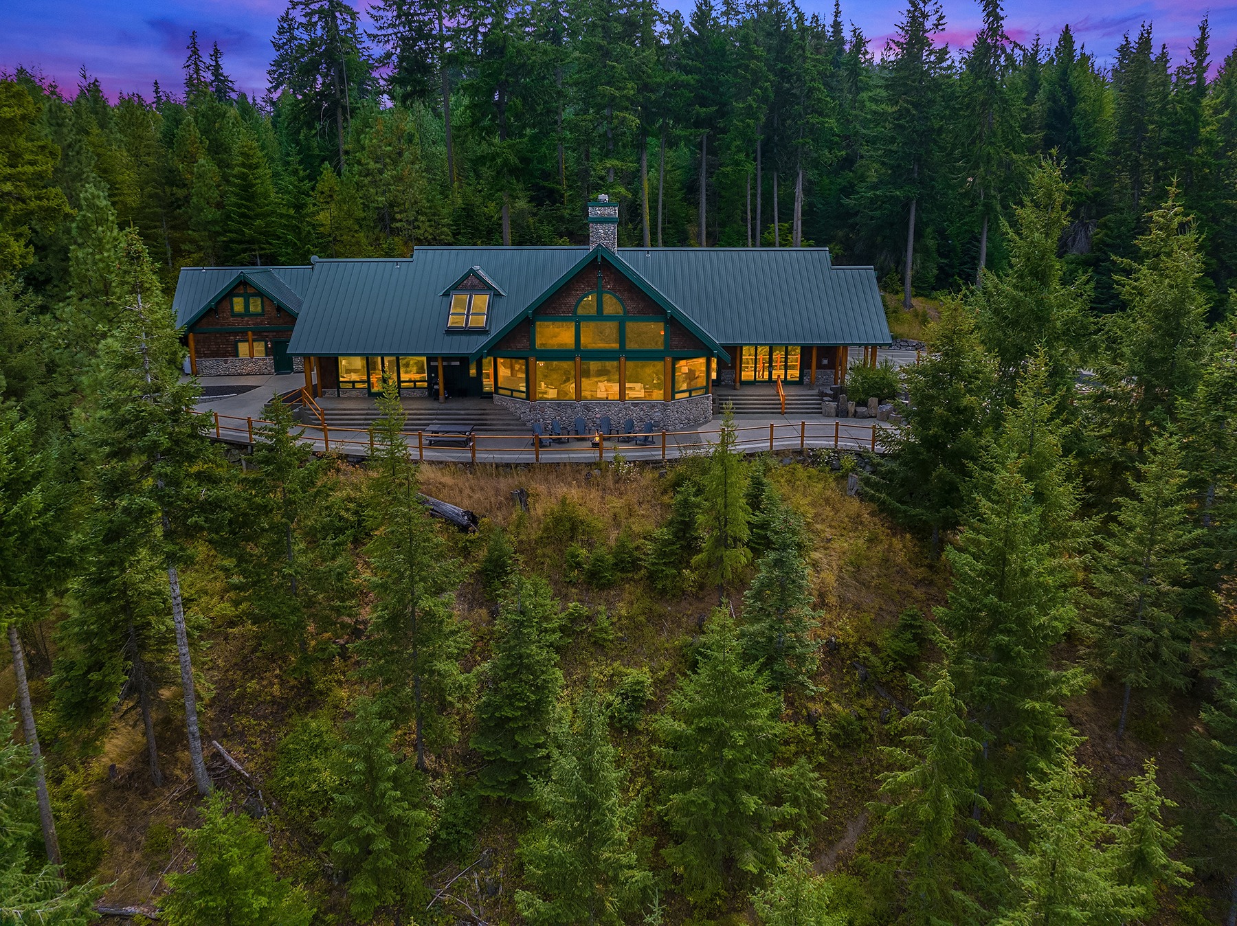 A large rustic cabin with a green roof is nestled amidst densely forested trees. The cabin has multiple large windows glowing warmly from inside. The scene is set under a twilight sky with a mix of purples and blues, suggesting sunset or dusk.