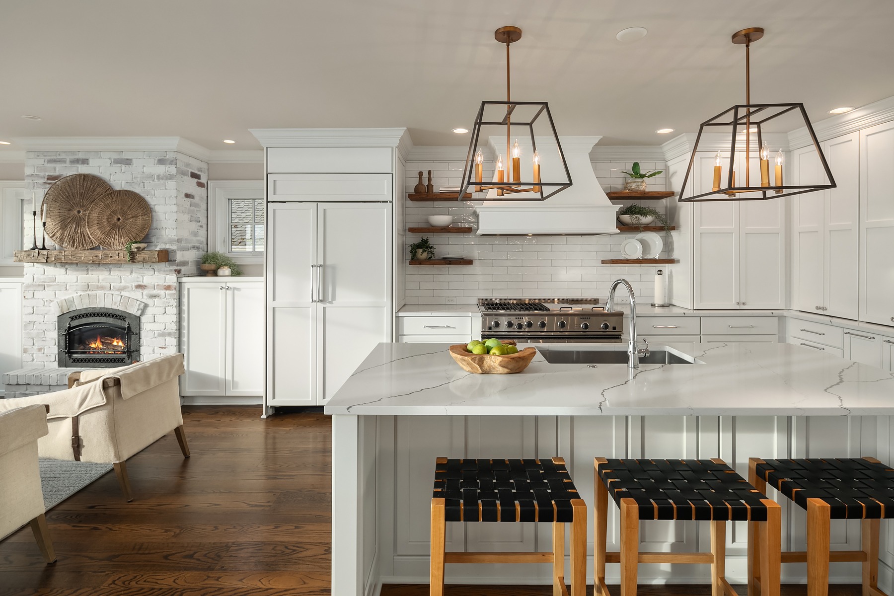 A modern kitchen with white cabinetry, marble countertops, and stainless steel appliances. Two geometric pendant lights hang above the island, which features woven barstools. A cozy sitting area with a fireplace is visible in the background.