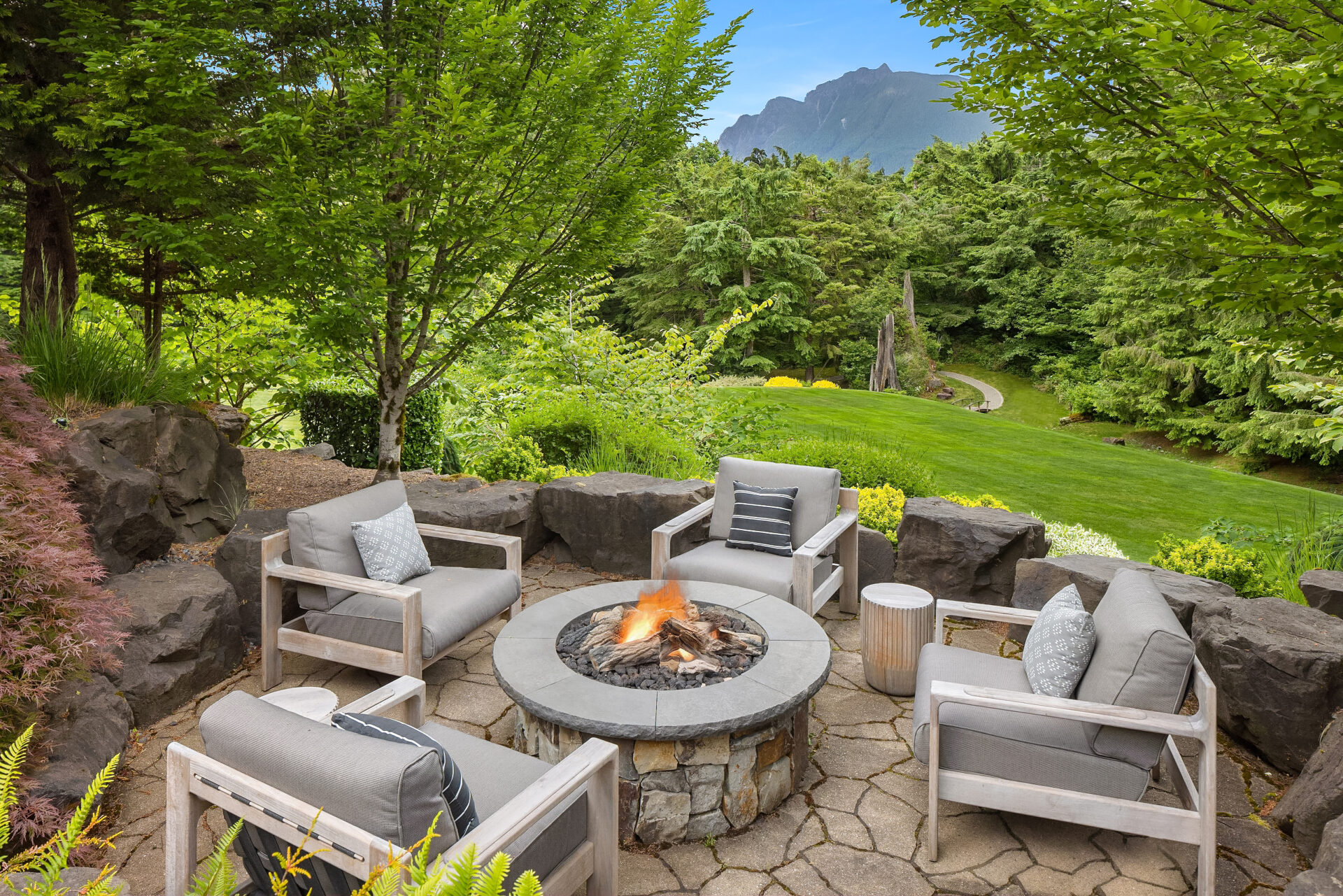 A circular stone fire pit is surrounded by four cushioned outdoor chairs on a patterned stone patio. The area is lush with green trees and plants, and a well-manicured lawn extends into the distance with mountains visible beyond the trees.