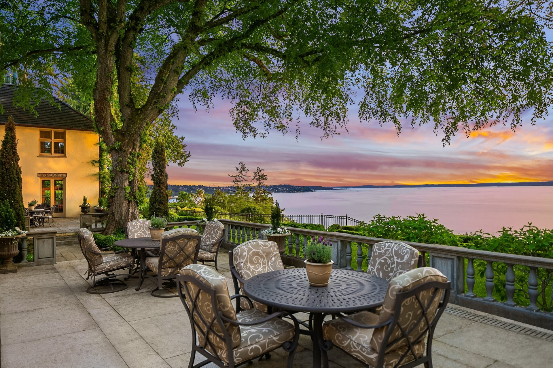 A scenic outdoor patio with elegant furniture under a large tree. The seating area overlooks a serene body of water with a stunning sunset sky filled with pink, orange, and purple hues. A charming house with light yellow walls is partially visible to the left.