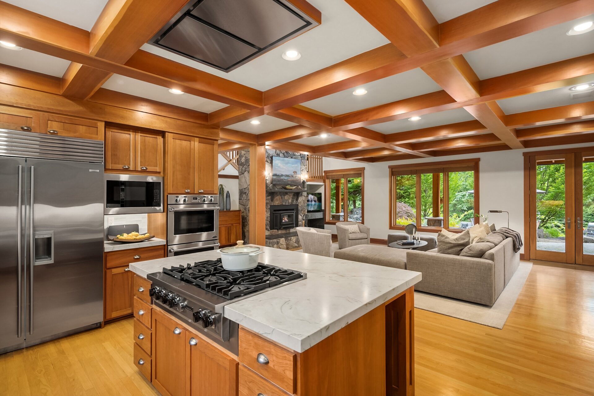 A modern open-concept kitchen and living area featuring a large kitchen island with a stovetop and marble countertop. Wooden cabinetry and stainless steel appliances adorn the kitchen. The living room has a gray sectional sofa and a stone fireplace.