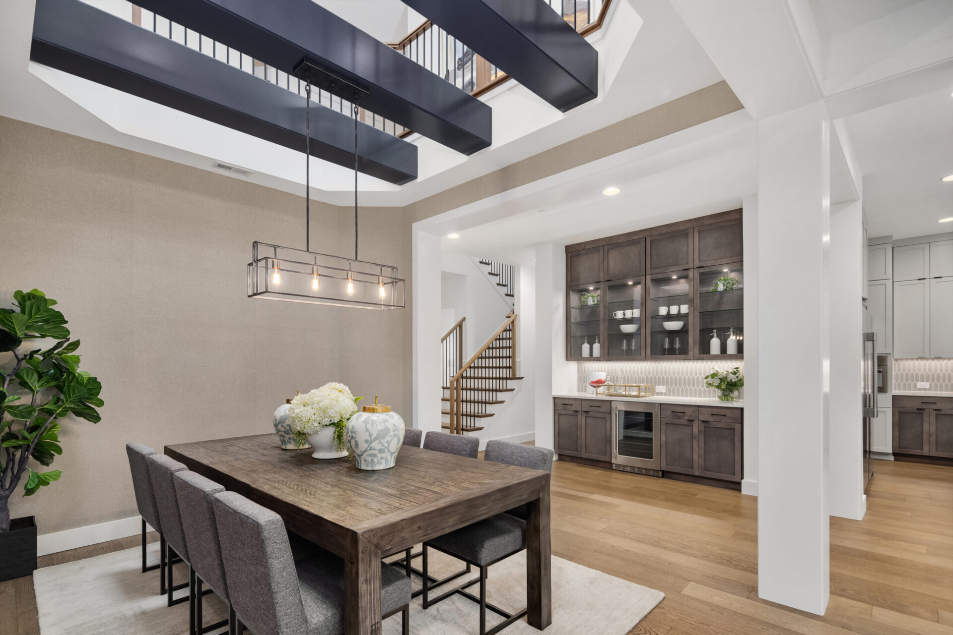 A modern dining room with a wooden table and six gray upholstered chairs. Above the table hangs a rectangular, linear chandelier with exposed bulbs. The background shows a staircase, built-in cabinets, and a plant in the corner, all under a high ceiling with beams.