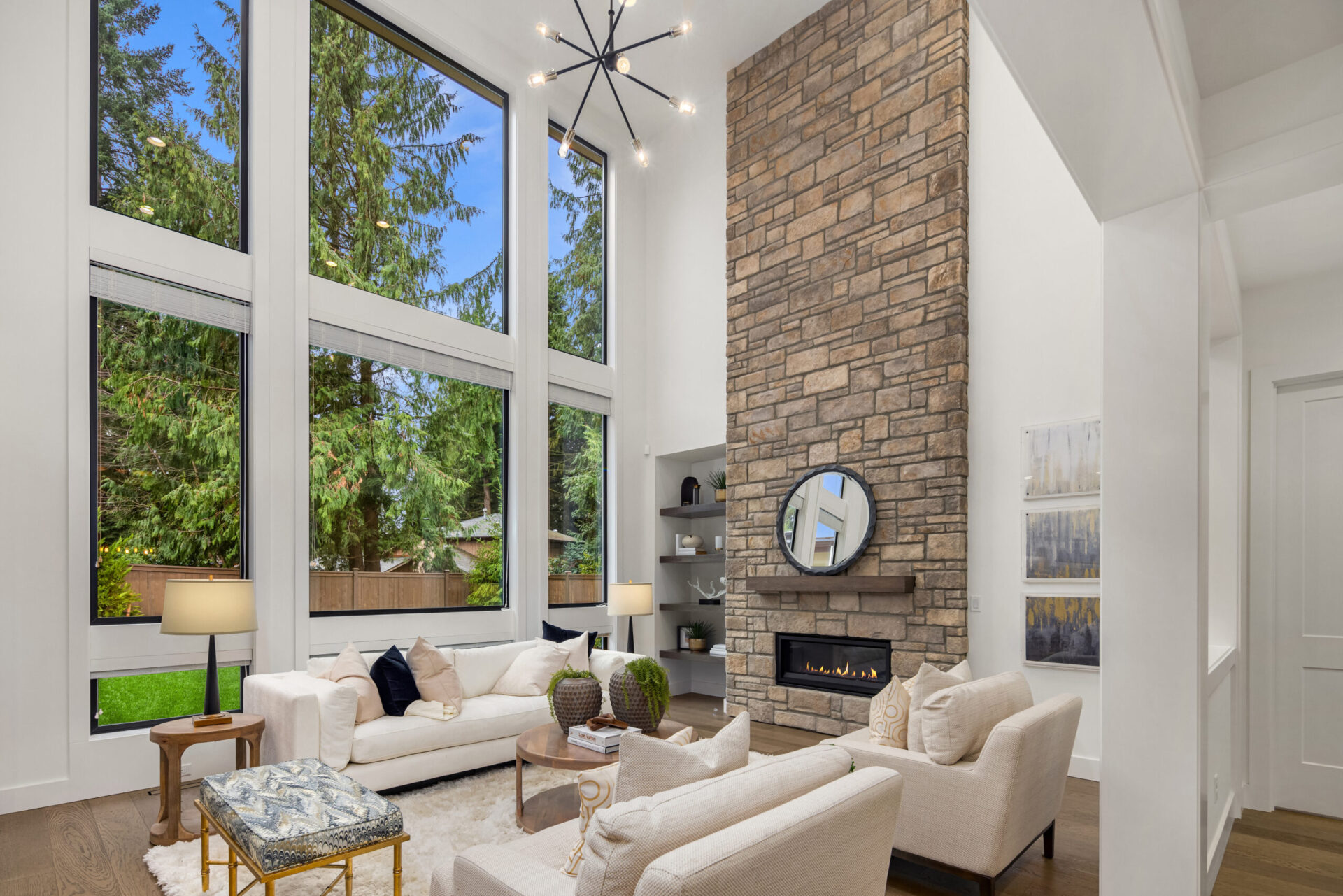 A modern living room with high ceilings, featuring a floor-to-ceiling stone fireplace topped with a round mirror. The space includes a white sofa, armchairs, and a coffee table. Tall windows offer a view of lush greenery outside. A contemporary light fixture hangs from the ceiling.