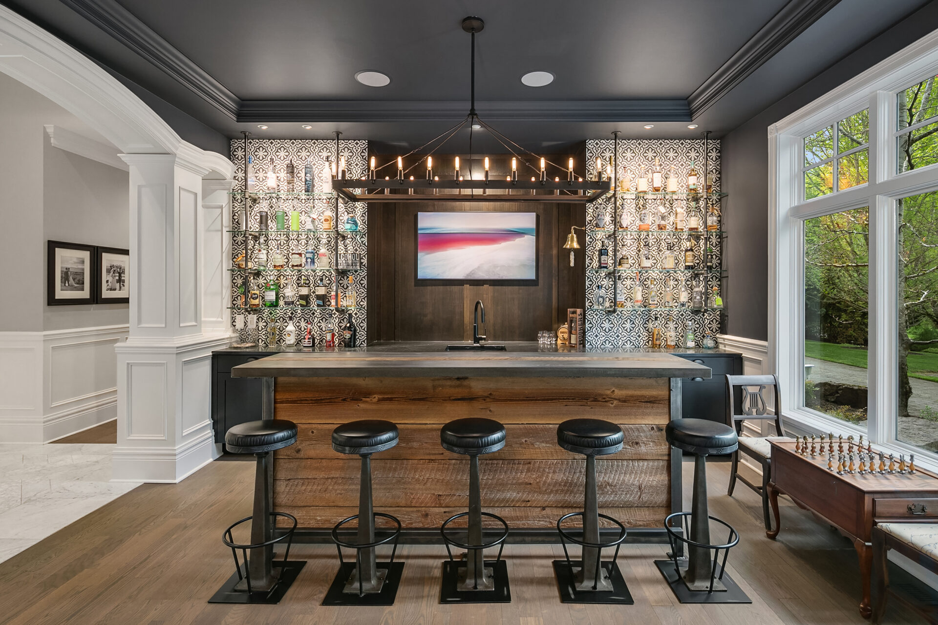 A modern home bar with a sleek wooden counter, four bar stools, and artistic black and white tile backsplash. Bottles are neatly arranged on backlit shelves. A colorful abstract painting hangs above the sink. Large windows offer views of lush greenery outside.