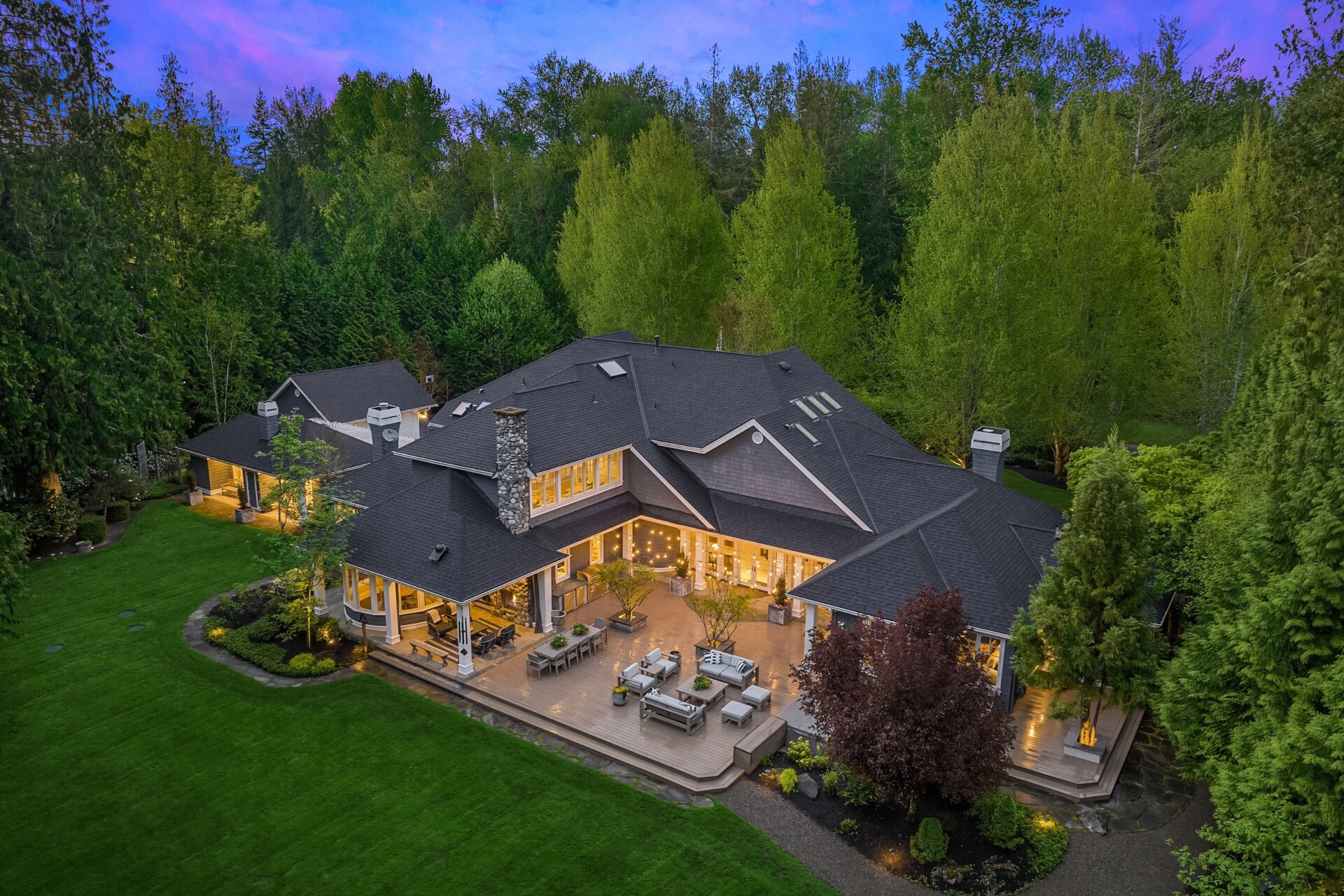 Aerial view of a spacious, modern house with a dark roof, surrounded by lush greenery and trees. The house features multiple large windows and an expansive outdoor patio area with seating and dining arrangements, illuminated by warm lights. A well-maintained lawn encircles the property.