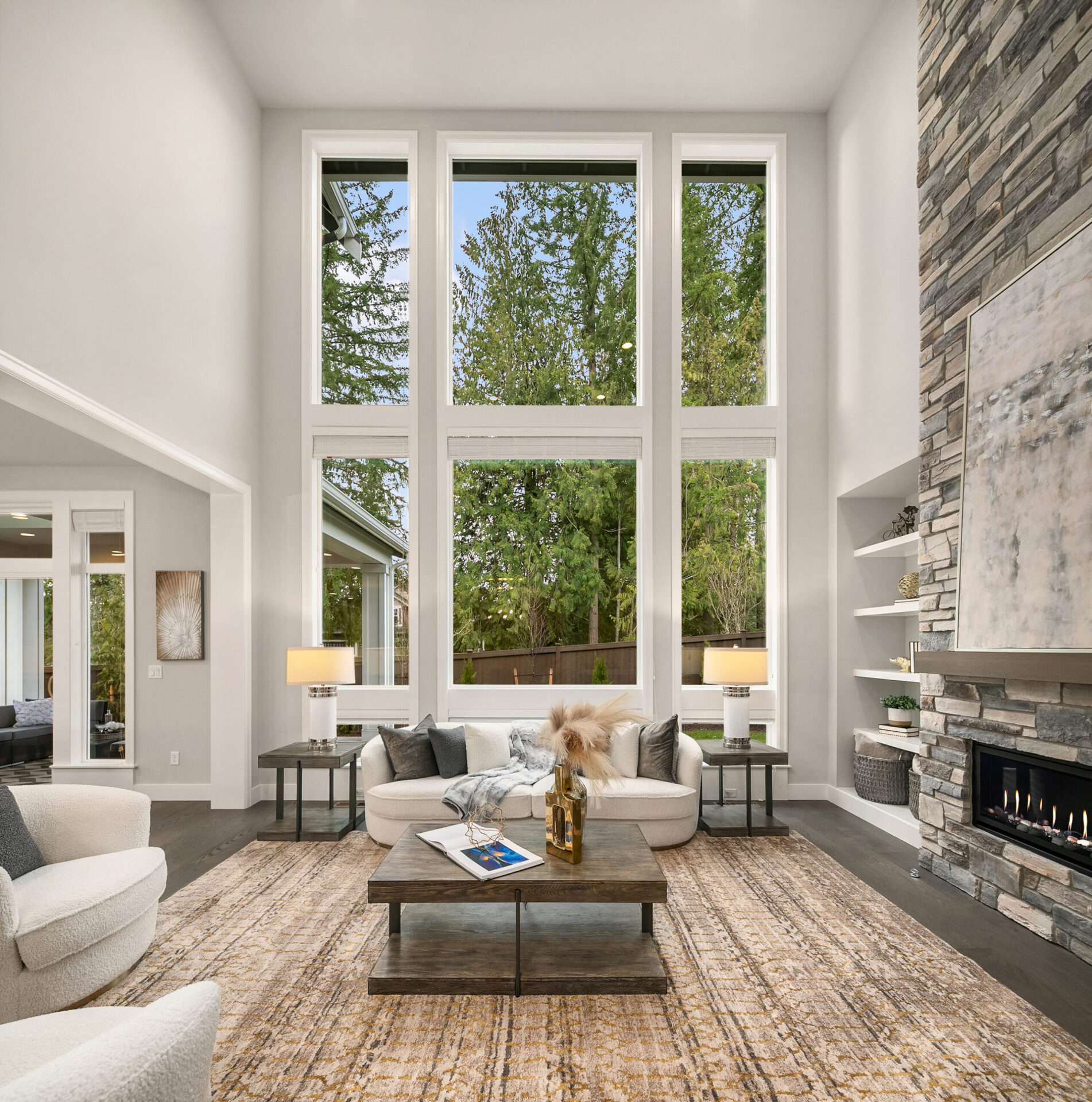 A spacious, modern living room with large floor-to-ceiling windows that offer a view of lush greenery. The room features a cozy stone fireplace, two white sofas, a rug, and a wooden coffee table with decorative items. Shelves with decor are built into the wall.