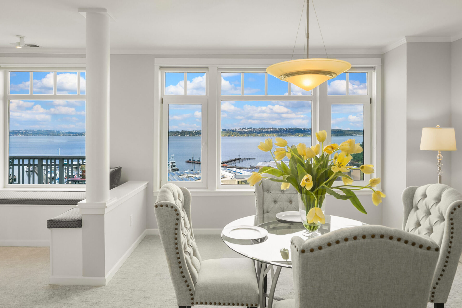 A dining area with a round glass table and four upholstered chairs. A vase with yellow flowers sits in the center. Large windows overlook a waterfront view with a bay and distant mountains. A cushioned bench under the window and a modern chandelier are also visible.