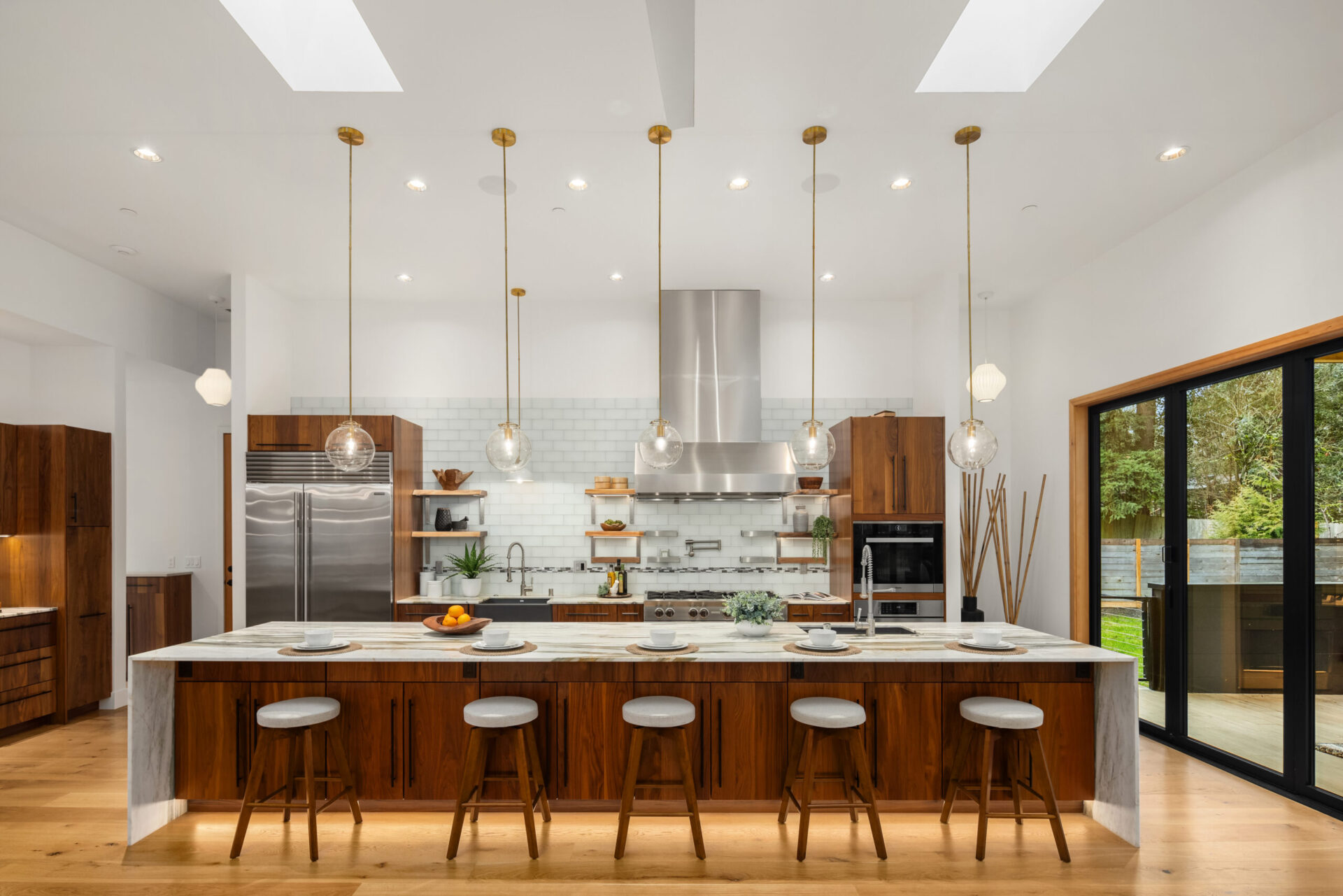 A modern kitchen with wooden cabinets and a large island. The island has bar stools and is topped with dishes and fruits. Pendant lights hang overhead. Stainless steel appliances, including a refrigerator and stove, and a tile backsplash are visible. Large windows bring in natural light.