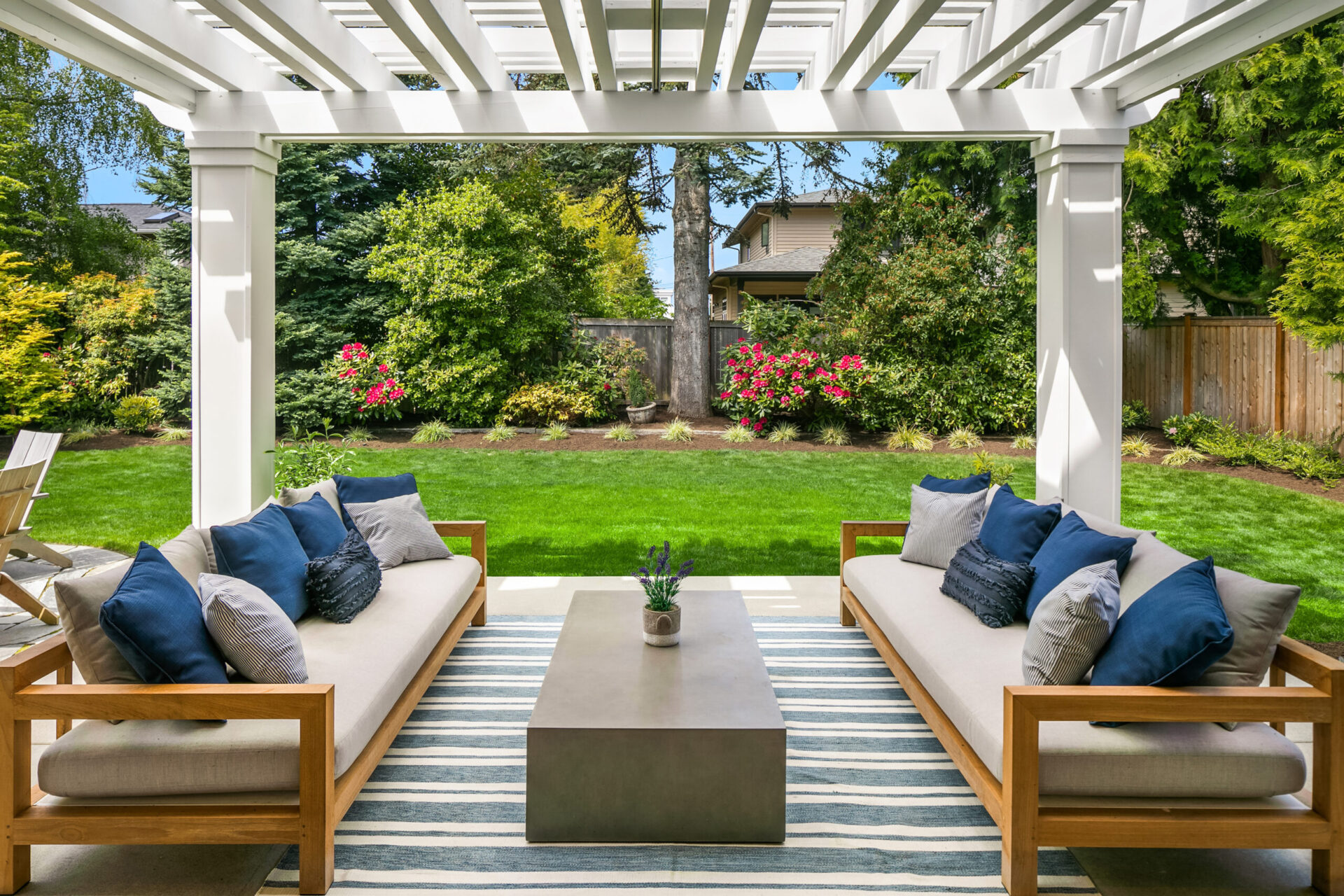 A patio with a white wooden pergola features two wooden sofas with beige cushions and blue throw pillows facing each other. A rectangular coffee table sits between them on a striped rug. Behind the patio, a lush green lawn and landscaped garden are visible.