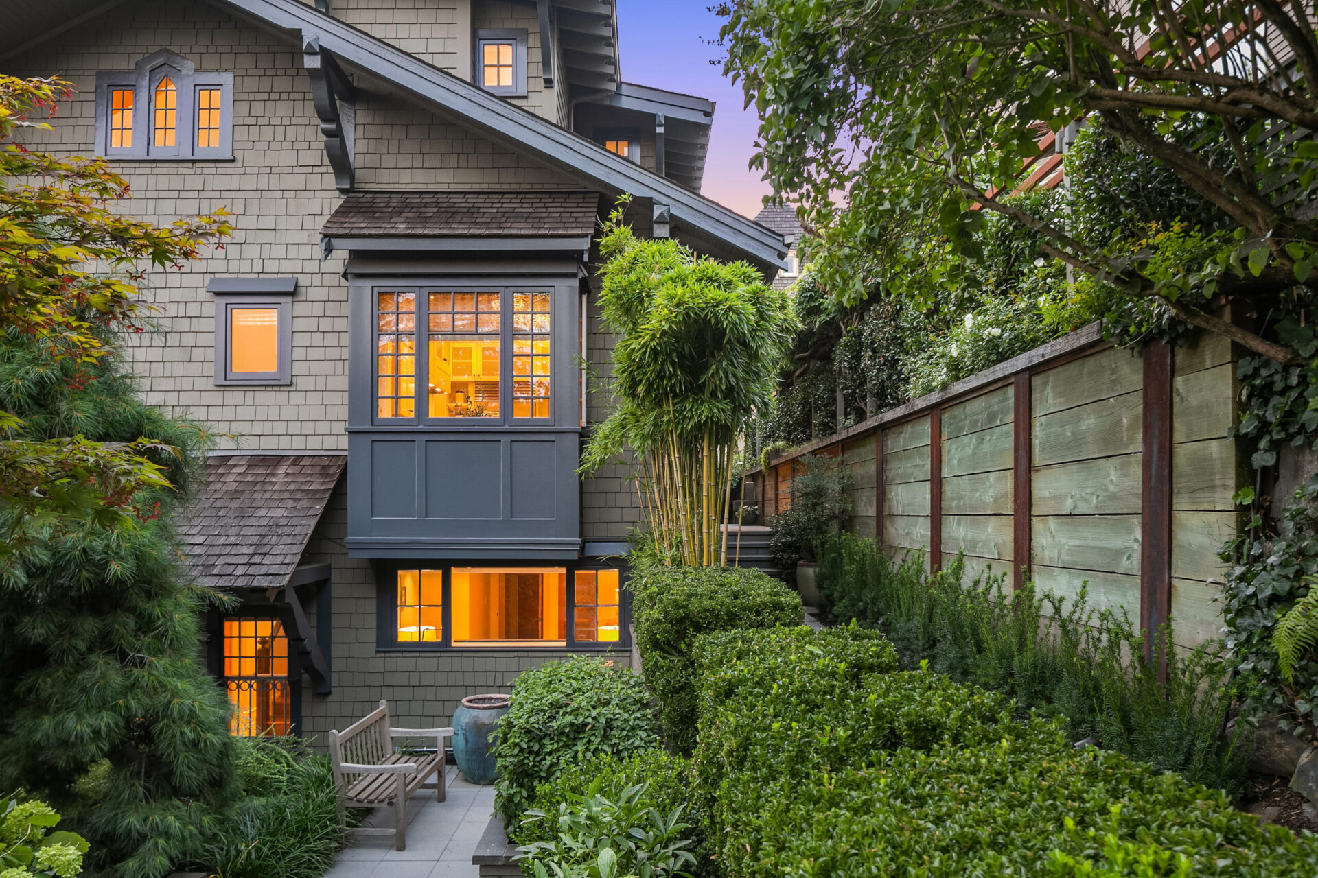 A charming multi-story house with wooden siding and large windows glowing with warm light. It features a lush, manicured garden with various green shrubs and plants, a wooden bench, and a tall wooden fence providing privacy. The sky is a soft twilight hue.