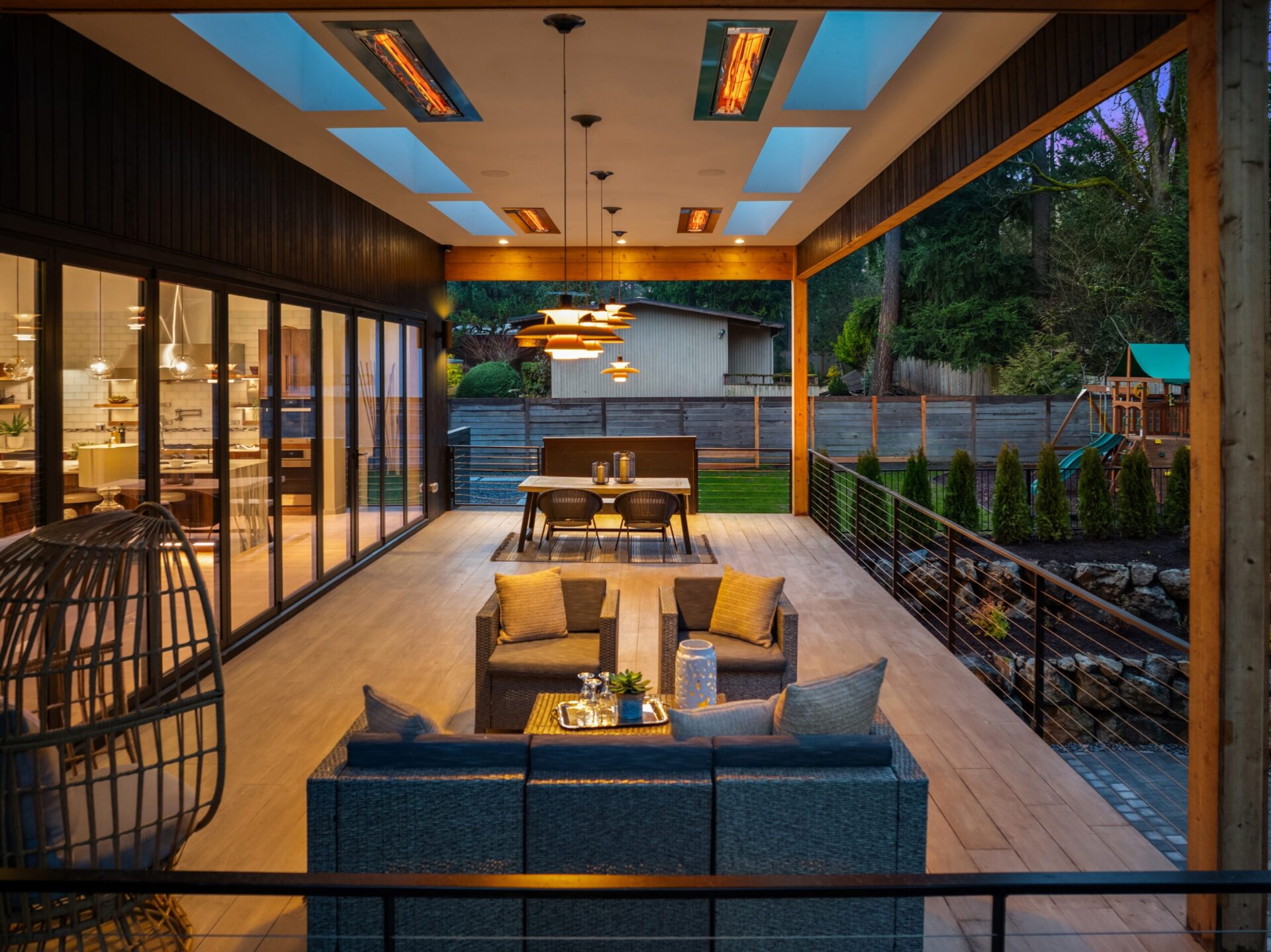 A modern outdoor patio with ceiling heaters and skylights. It features a lounging area with a gray wicker sectional and a hanging chair, a dining table, and an outdoor kitchen. Large sliding glass doors lead to the indoor area. A backyard with a playset is visible.