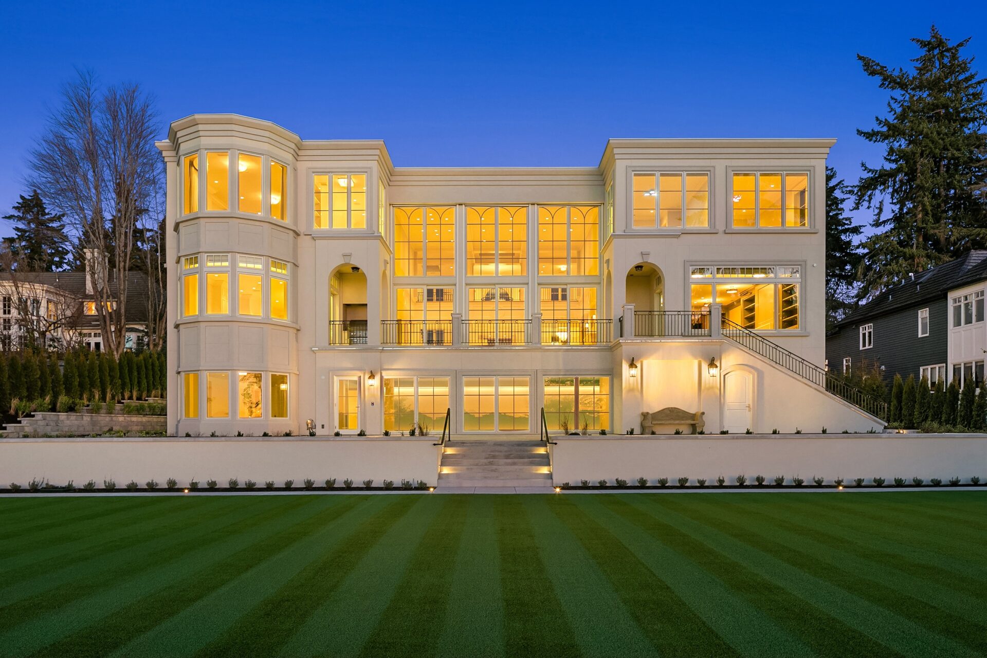 A large, elegant mansion with extensive windows is illuminated warmly at dusk. Its symmetrical design is highlighted by multiple balconies and a central staircase leading down to a meticulously manicured lawn. Trees and neighboring houses are visible in the background.