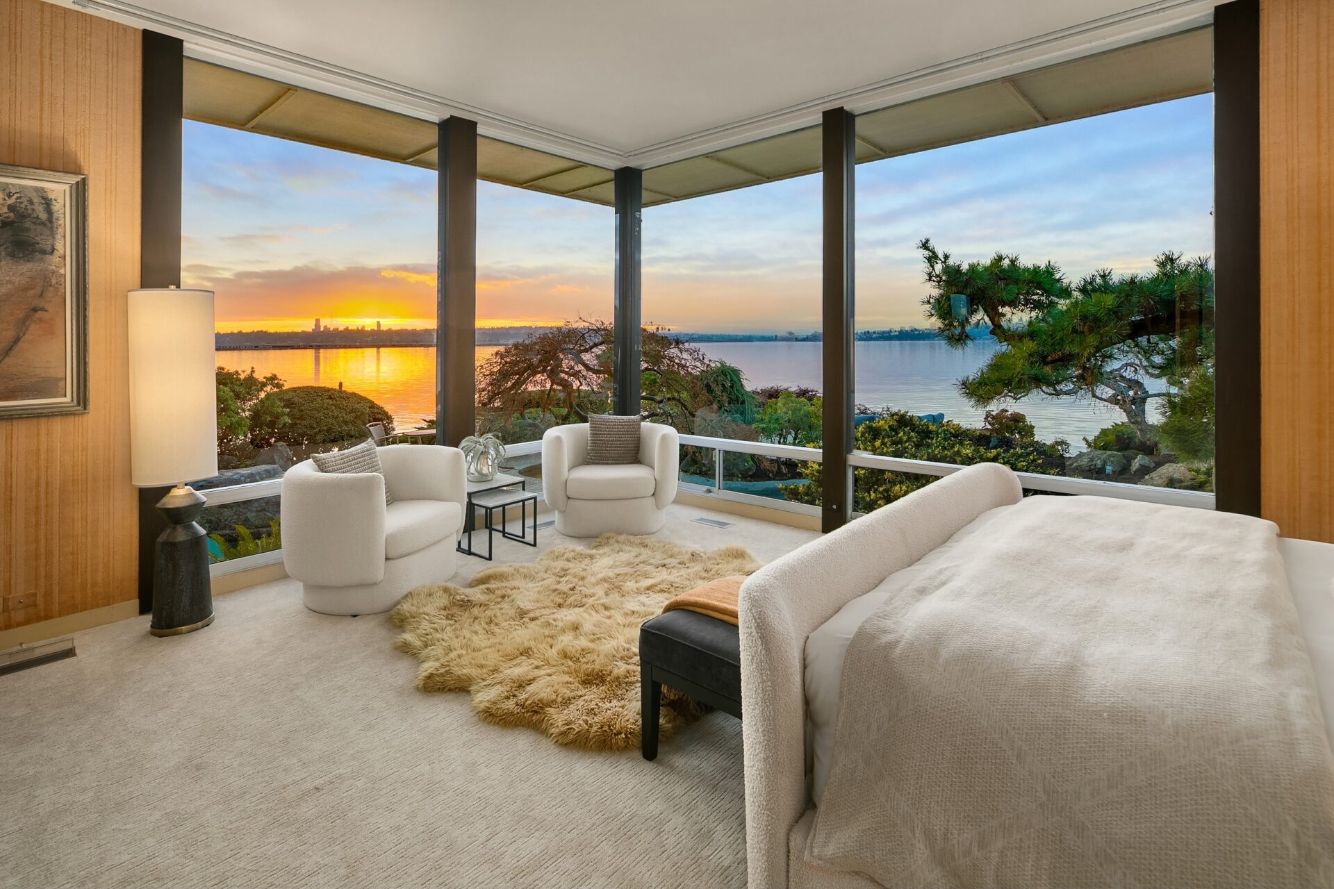 A modern bedroom with floor-to-ceiling windows offers a stunning view of a sunset over a body of water. The room is furnished with a bed, two white armchairs, a bench, and a fluffy rug on a light carpet. A lamp and decorative items are placed near the window.