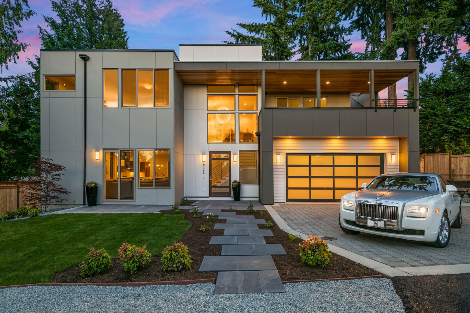 A modern two-story house with large windows, a flat roof, and a spacious balcony. It features a well-kept garden with stone walkways leading to the entrance. A luxury car is parked in the driveway beside the three-car garage. The sky has a pinkish hue at dusk.