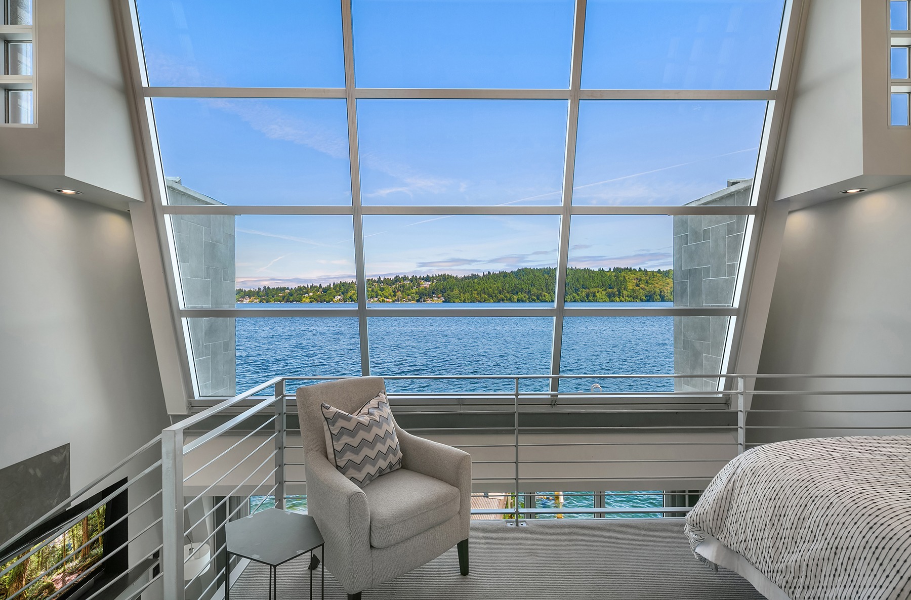 A modern bedroom with a large, slanted floor-to-ceiling window that offers a scenic view of a blue lake and a green forested area on the other side. The room features a gray armchair, a small side table, and a part of a bed with a white comforter.