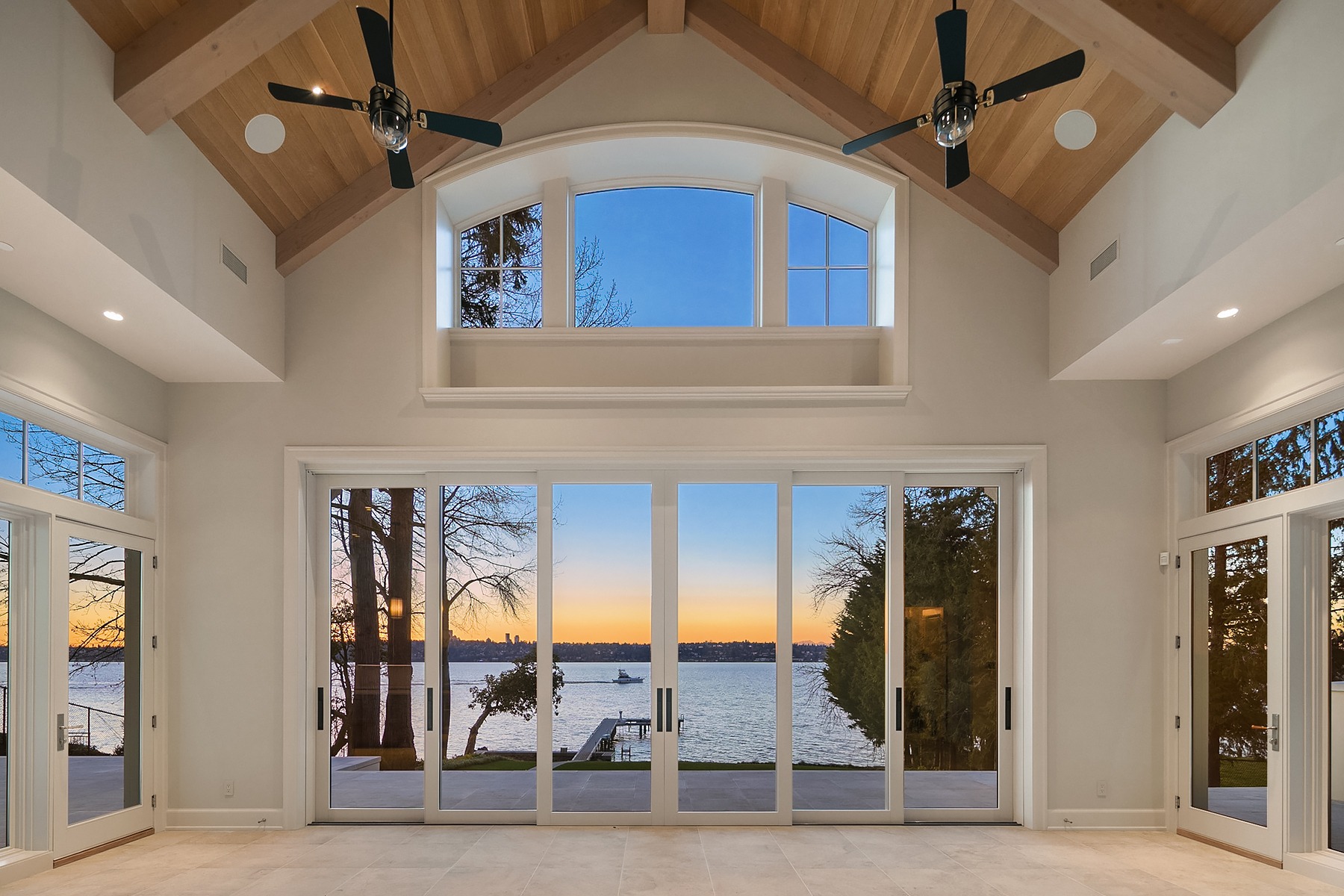 A spacious room features a high vaulted wooden ceiling with two ceiling fans. Large windows and glass doors offer a panoramic view of a serene lake at sunset, framed by trees. The warm glow of the sunset creates a tranquil and inviting atmosphere inside.