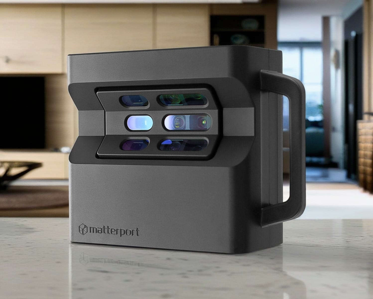 A black, box-shaped Matterport camera with multiple lenses sits on a marble countertop in a modern, bright living room. The camera has a handle on the right side and is used for capturing 3D images of spaces. The background features wooden cabinetry and a window.