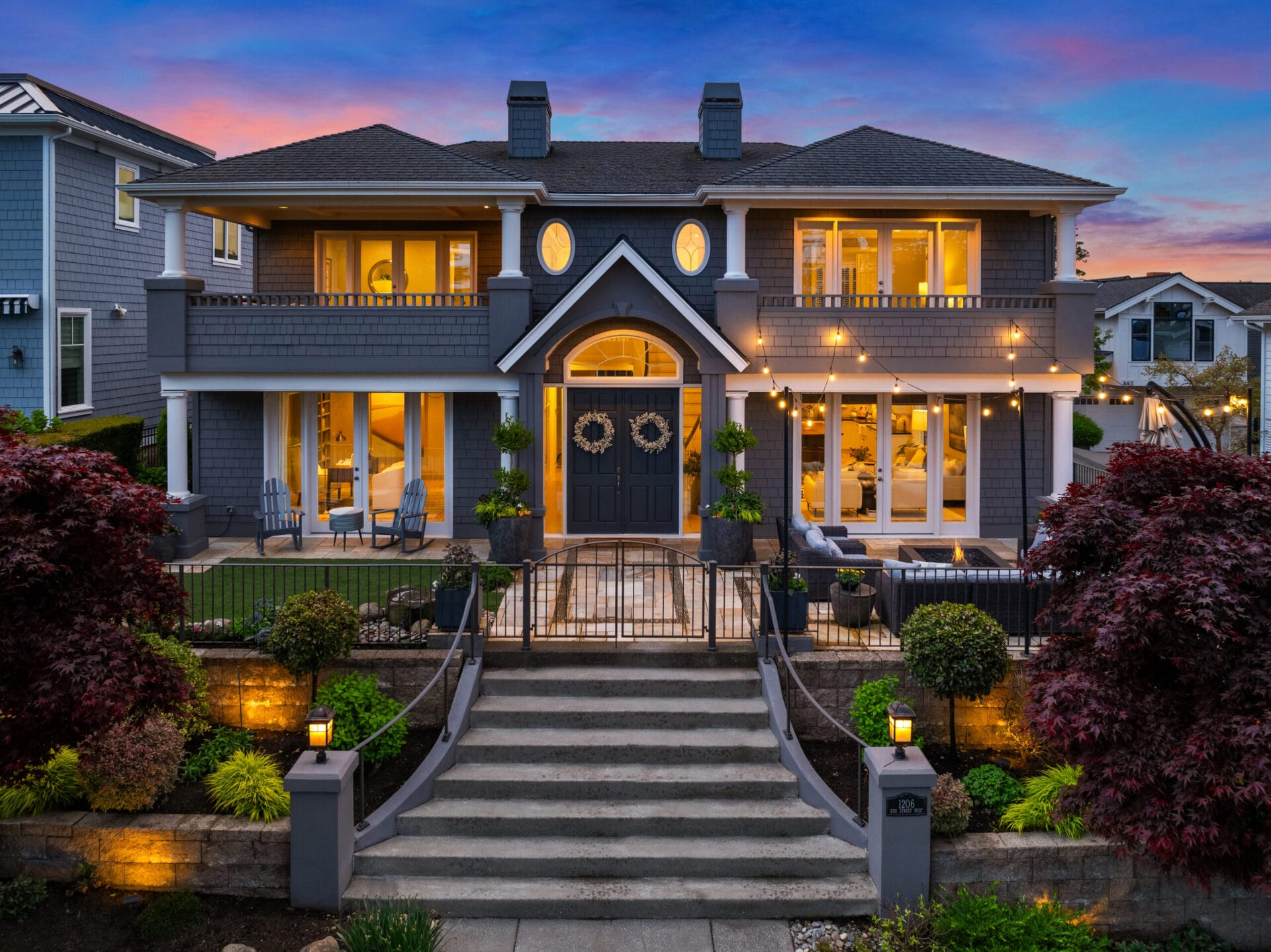 A two-story, grey suburban home with symmetrical windows and double front doors, warmly lit at twilight. The house features a front porch, a central staircase leading up to the entrance, manicured landscaping, and string lights hanging above the patio.