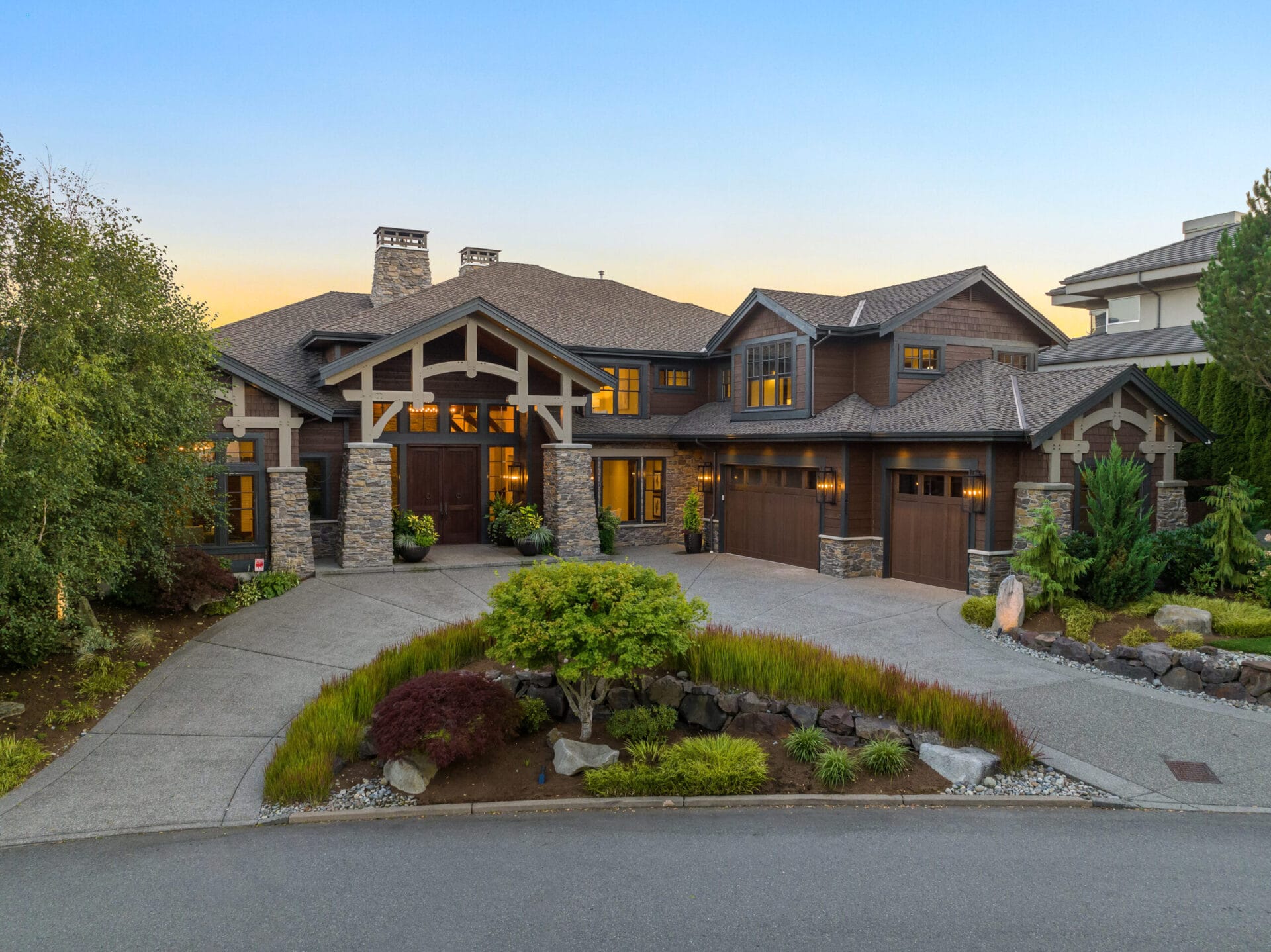 A large, contemporary house with a stone and wood exterior, featuring a blend of rustic and modern design elements. The driveway leads to a three-car garage, and the landscaped front yard is adorned with shrubs, trees, and a small rock garden. The sky is clear at dusk.