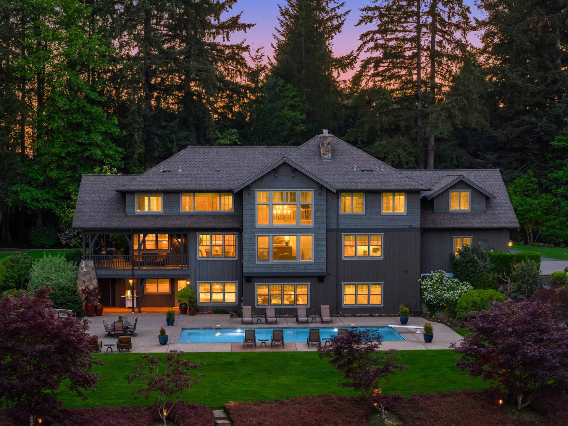 A large, modern house with numerous illuminated windows is surrounded by tall trees. In the backyard, there's a spacious patio area with a swimming pool and lounge chairs. The sky is beautifully lit by the colors of the setting sun.