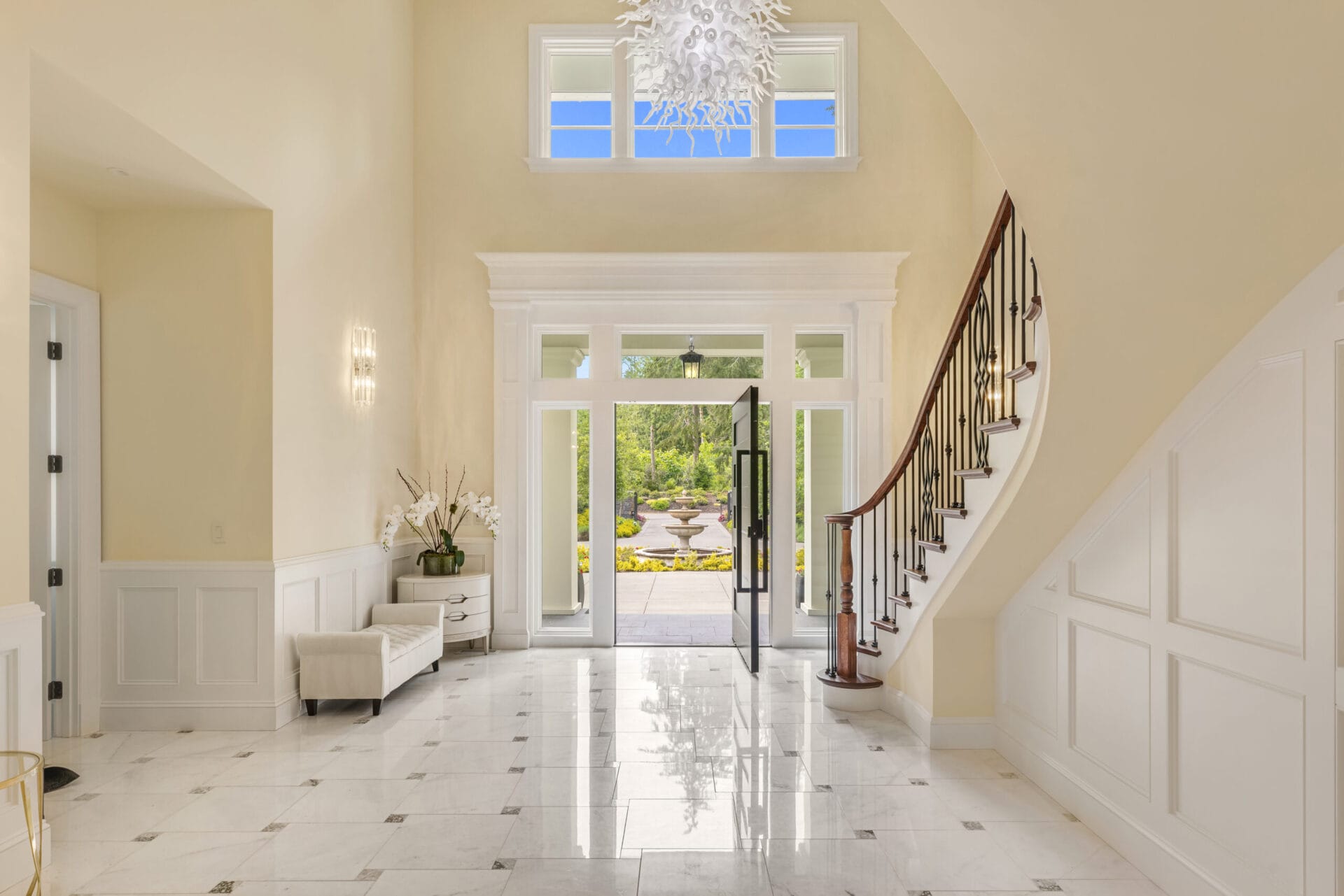 A spacious and elegant foyer with a grand staircase curving to the right. The floor is tiled in white with subtle accents. Large windows above and beside the entrance door flood the room with natural light. A modern crystal chandelier hangs from the ceiling.