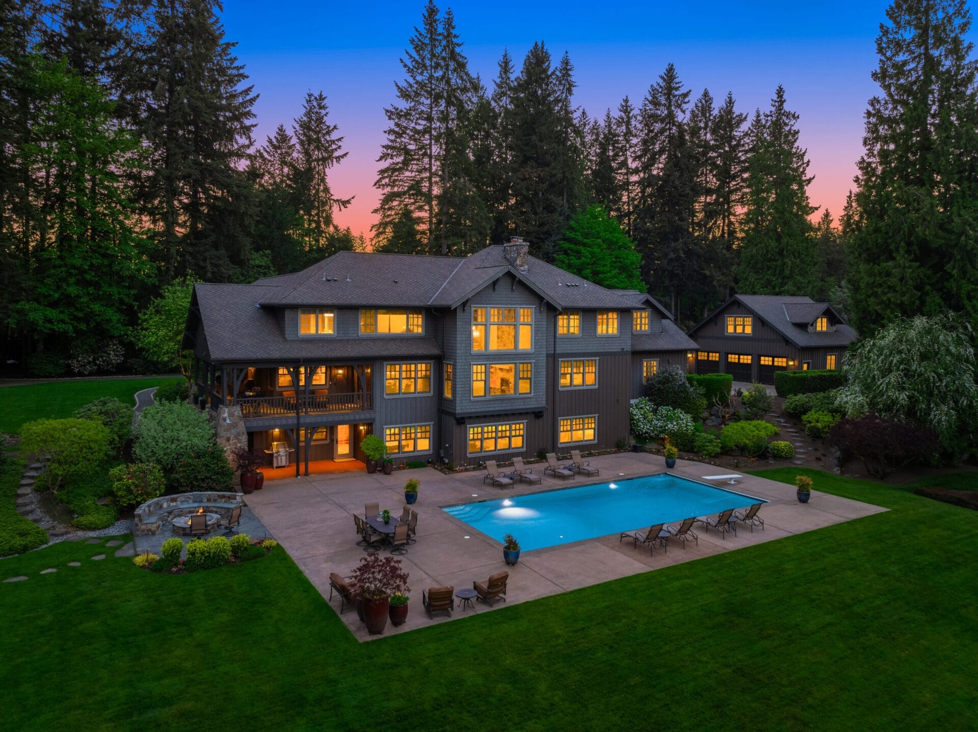 A large, multi-story house with numerous lit windows is surrounded by tall trees at dusk. The backyard features a spacious patio, swimming pool, lounge chairs, and manicured lawn. Additional buildings are visible in the background.