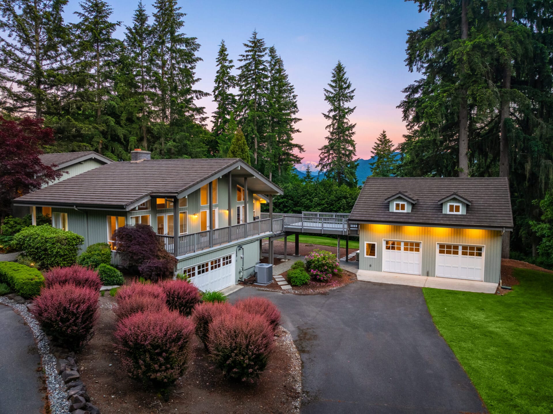 A beautiful two-story house with large windows, surrounded by lush greenery and tall trees. The home features an attached garage and a separate garage to the right. The property has a landscaped yard with trimmed bushes and a paved driveway. The sky is at sunset.