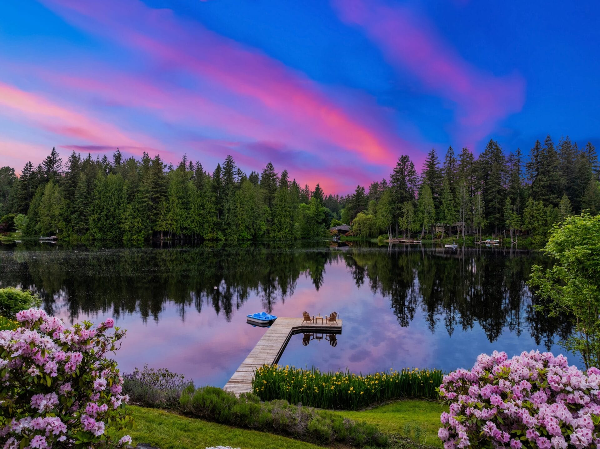 A serene lake at sunrise with a dock extending into the water, featuring a small boat and a couple of deck chairs at the end. Surrounding the lake are lush trees, vibrant flowers in the foreground, and a sky painted with stunning pink and purple hues.