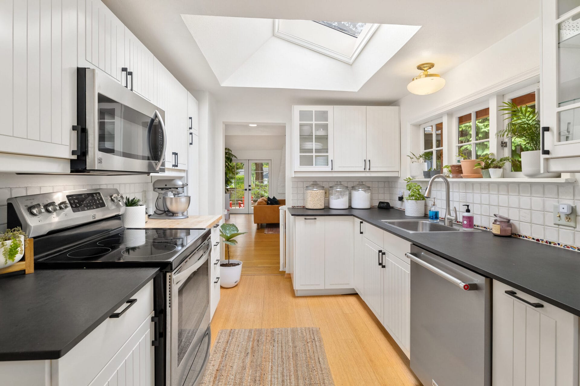 A modern, bright kitchen featuring white cabinetry, black countertops, and stainless steel appliances, including a stove and microwave. A large skylight and window bring in natural light, illuminating the space and potted plants. A woven rug lies on wooden flooring.