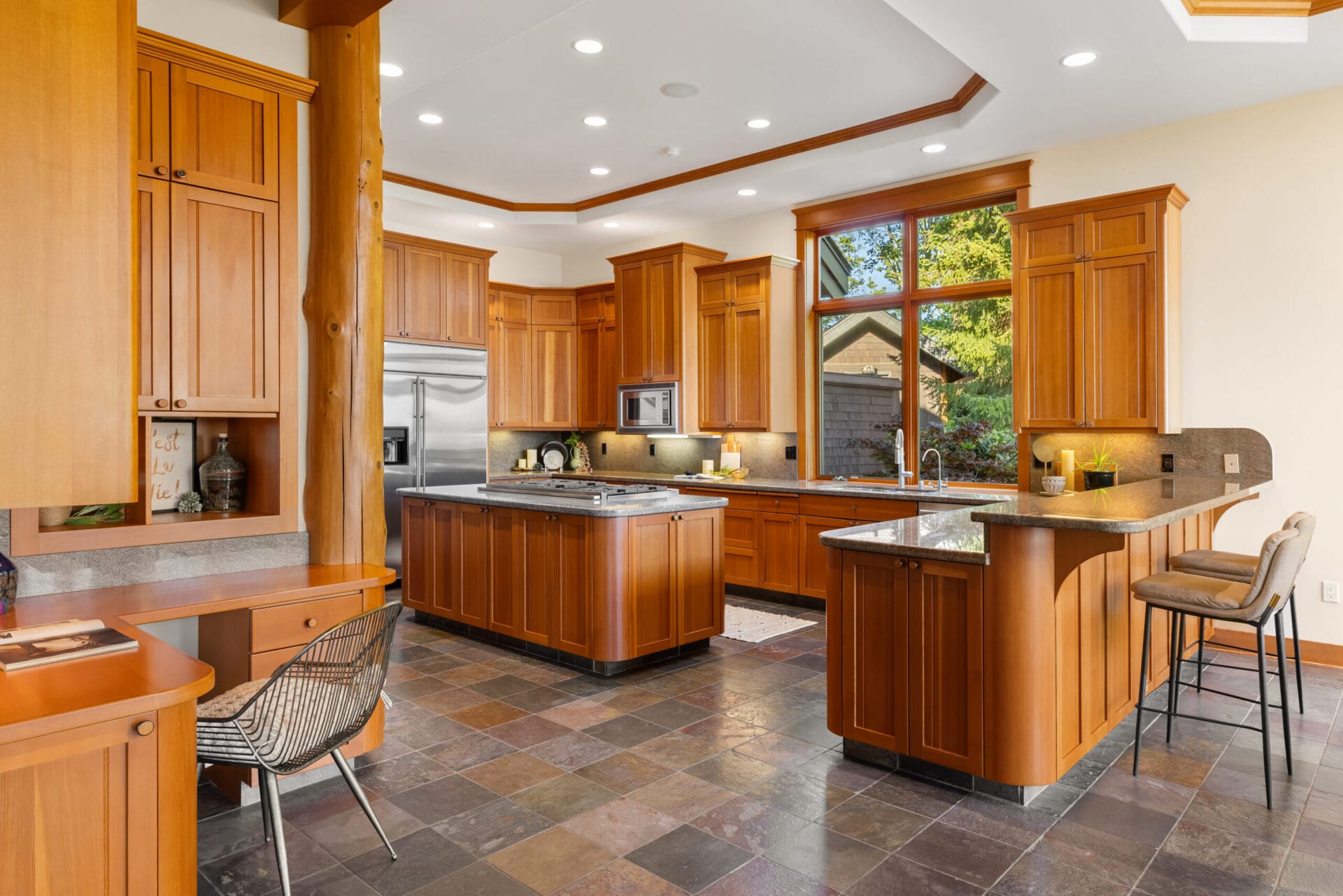 A spacious kitchen with wooden cabinetry, a central island, and modern stainless steel appliances. Large windows bring in natural light, highlighting the slate tile floor. There's a small desk area with a wicker chair on the left and a breakfast bar with stools on the right.