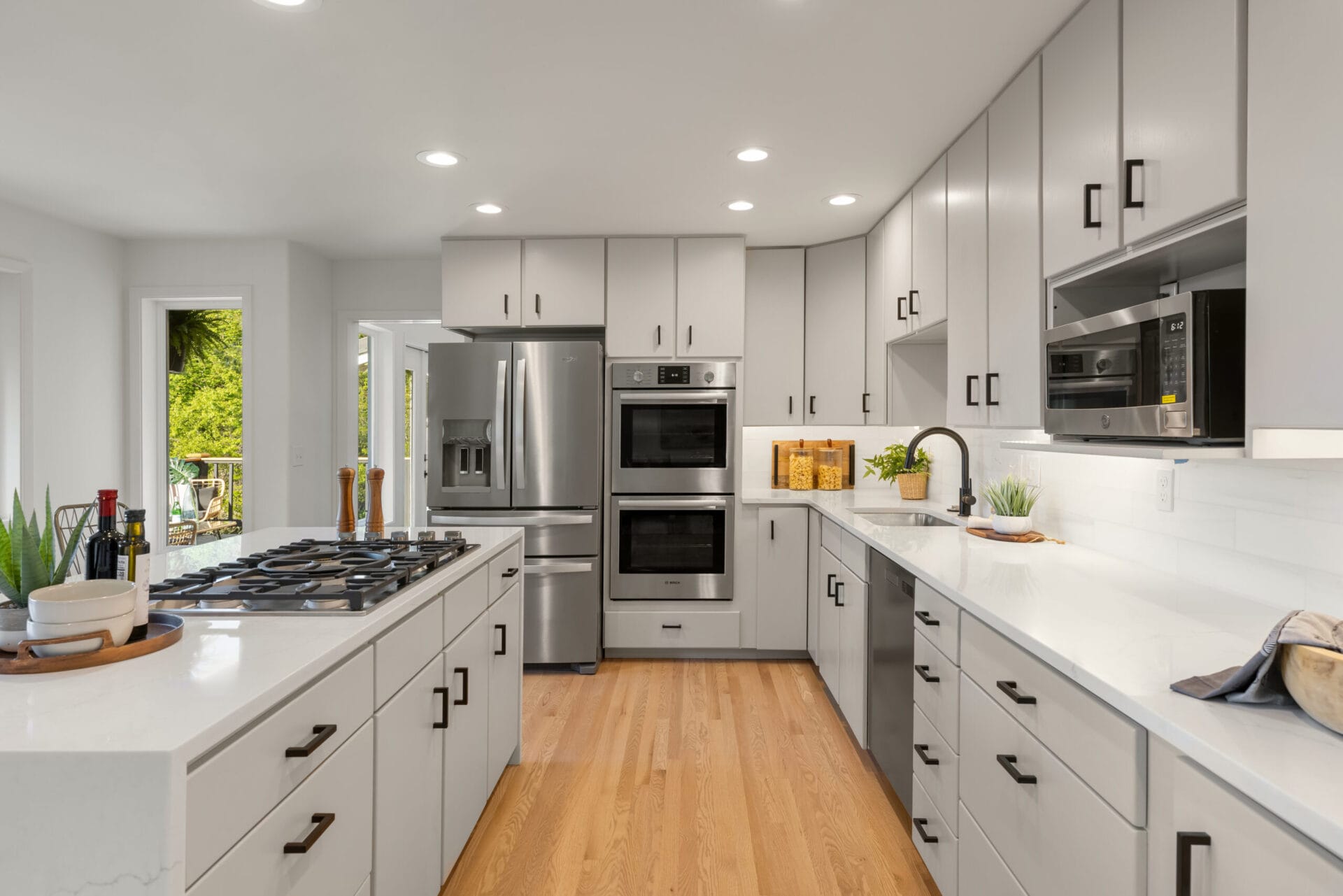 A modern kitchen with light gray cabinets, white countertops, and stainless steel appliances, including a refrigerator, oven, and microwave. The space has wood flooring, a gas cooktop on an island, and various decor items and kitchen essentials.