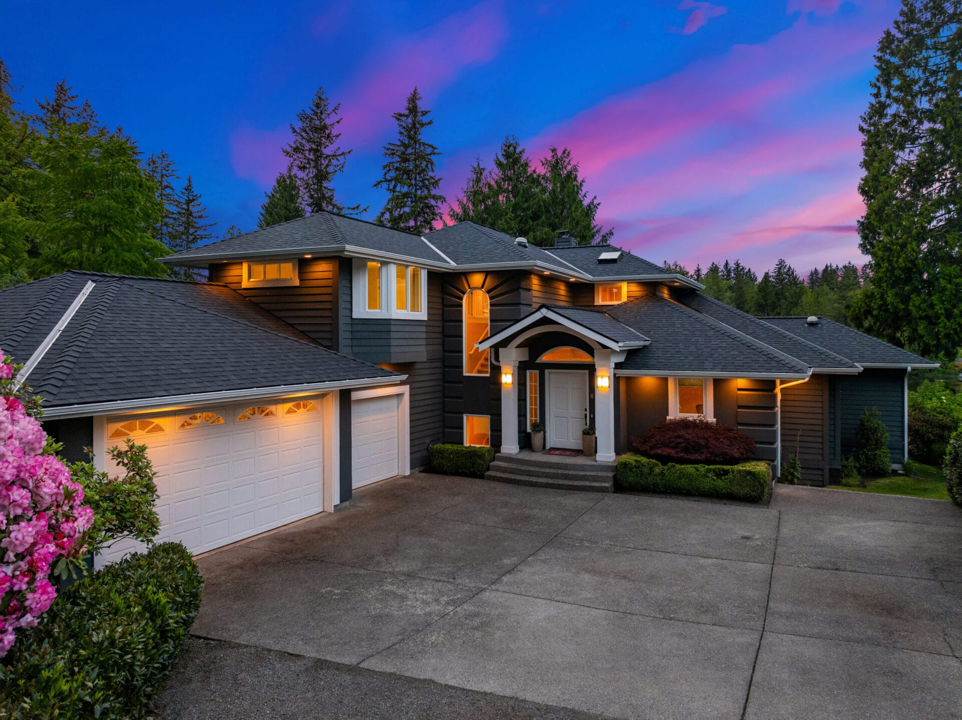 A spacious two-story house with gray siding and a white front door is surrounded by landscaped greenery. The house features a large driveway leading to a two-car garage. The sky is a vibrant mix of pinks and purples during sunset, with tall trees in the background.