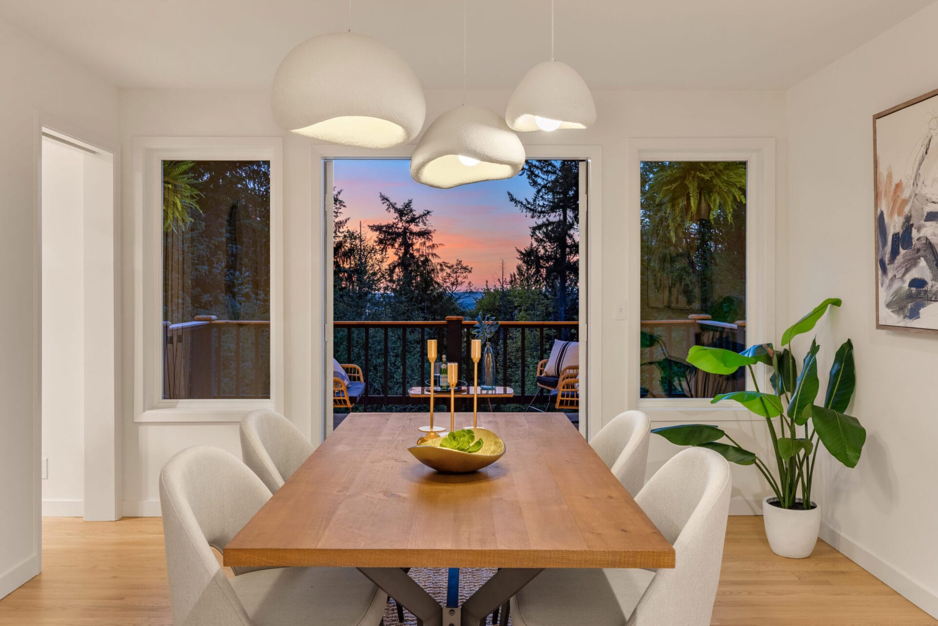 A dining room with a wooden table and six white upholstered chairs. Three pendant lights hang above the table. A large bowl of green apples is at the center of the table. There are two large windows and a door leading to a balcony with a sunset view. A plant stands in the corner.