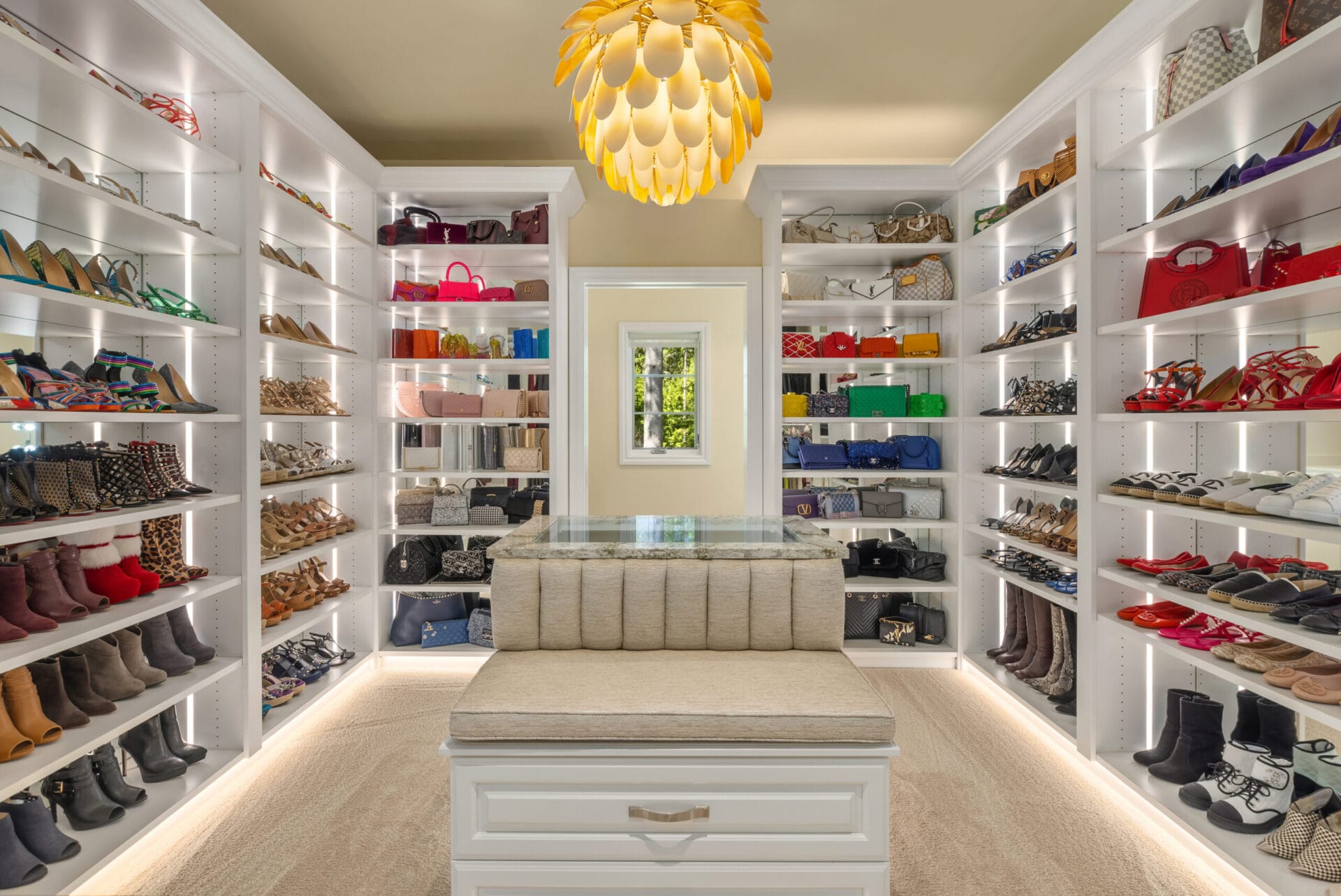 A luxurious walk-in closet featuring illuminated shelves displaying a collection of high-end shoes, handbags, and accessories. A stylish cushioned bench sits in the center of the room under an elegant gold chandelier. A mirror is positioned at the back wall.