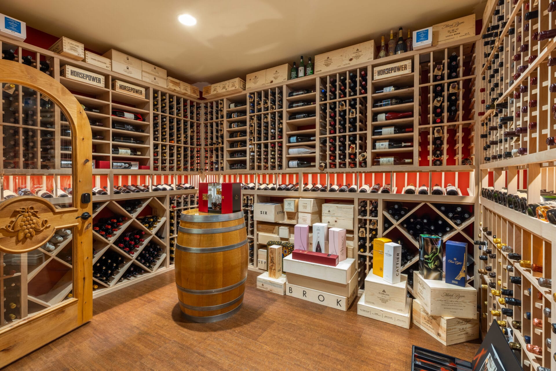 A well-organized wine cellar featuring wooden racks filled with various bottles of wine, a decorative wooden door with an oval glass insert, and a round wooden barrel placed centrally in the room. Boxes of wine and wine accessories are stacked against the walls.