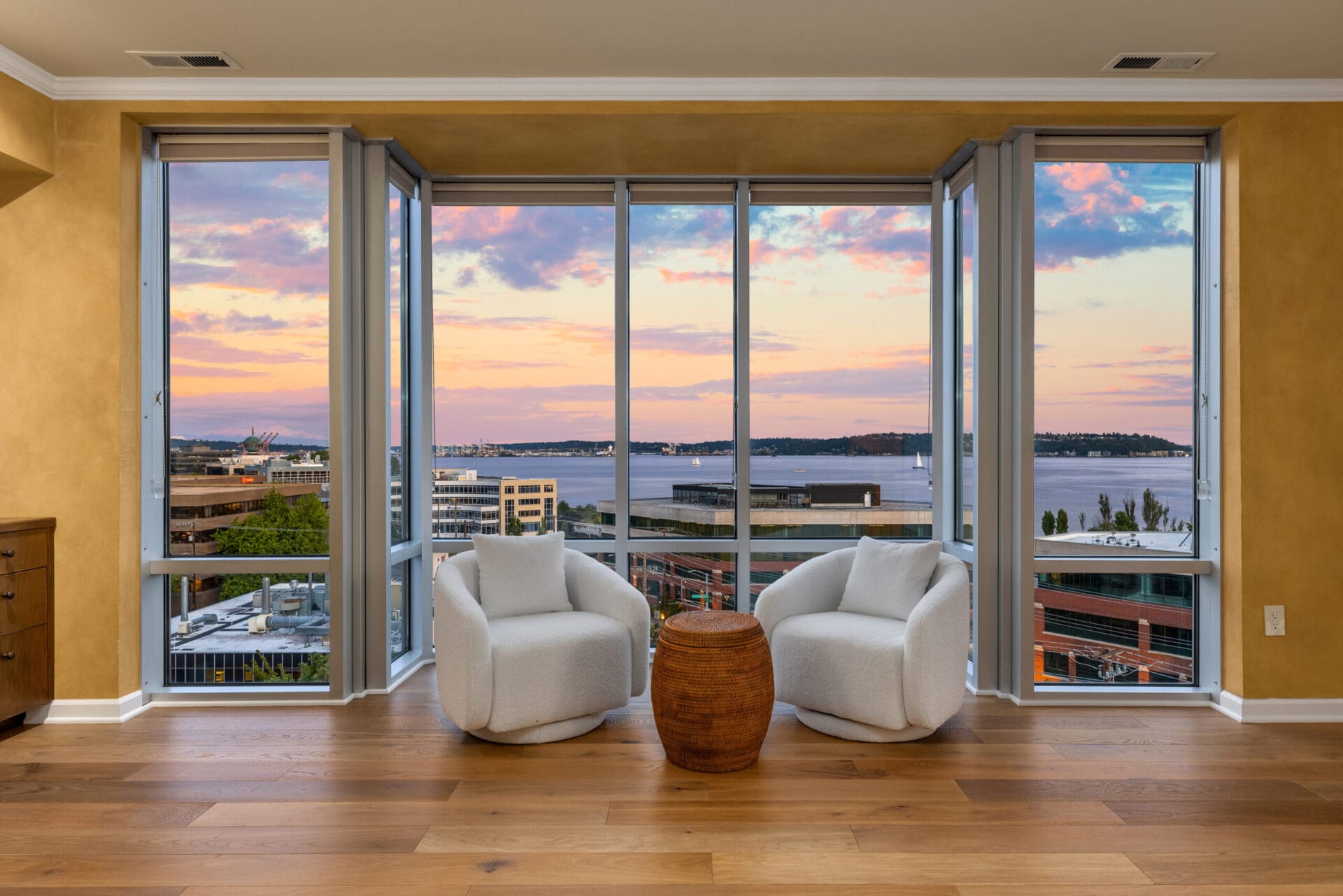 A well-lit room with large floor-to-ceiling windows offers a stunning view of a sunset over a body of water and cityscape. Two modern white chairs and a small wooden table are positioned in front of the windows on a hardwood floor.