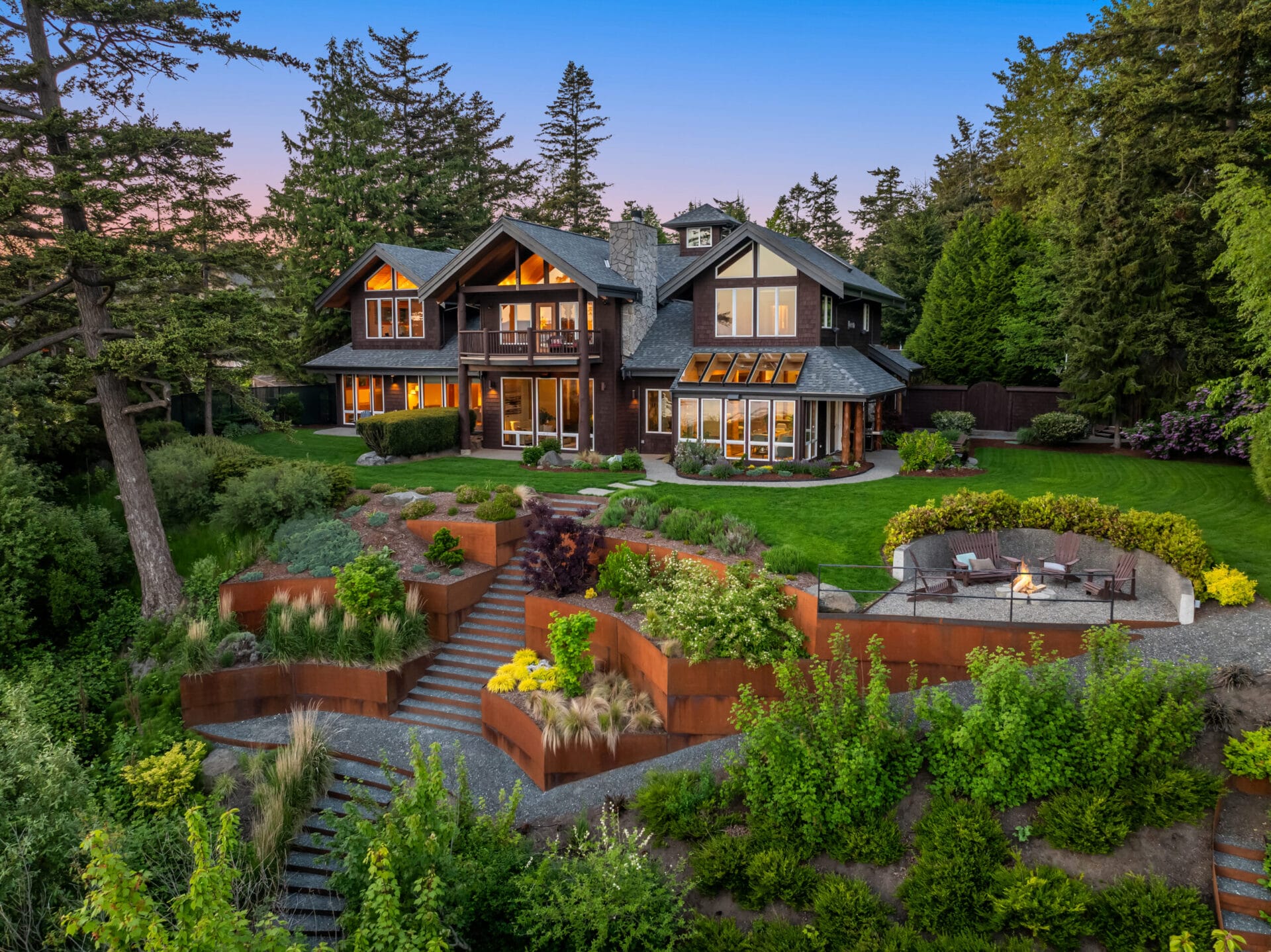 A luxurious two-story house with large windows and an intricate garden landscape featuring multiple levels, stone pathways, and vibrant plants. The backyard has a cozy fire pit area with seating. The scenery includes tall trees and a twilight sky.