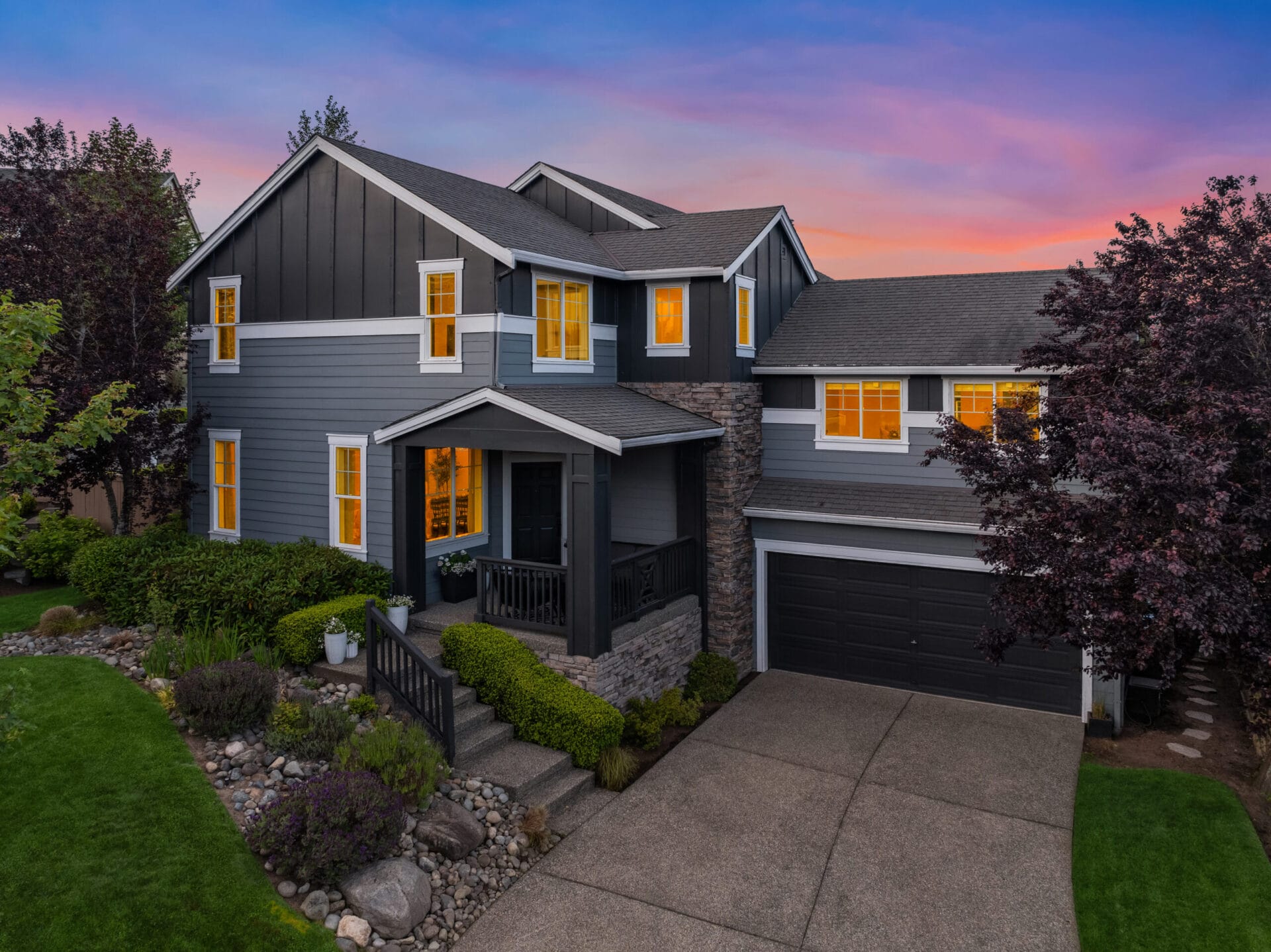 A two-story house with dark gray siding and white trim is illuminated by interior lights at dusk. The front yard features landscaped plants, stones, and a well-maintained lawn. The house has a front porch, a two-car garage, and trees framing the property.