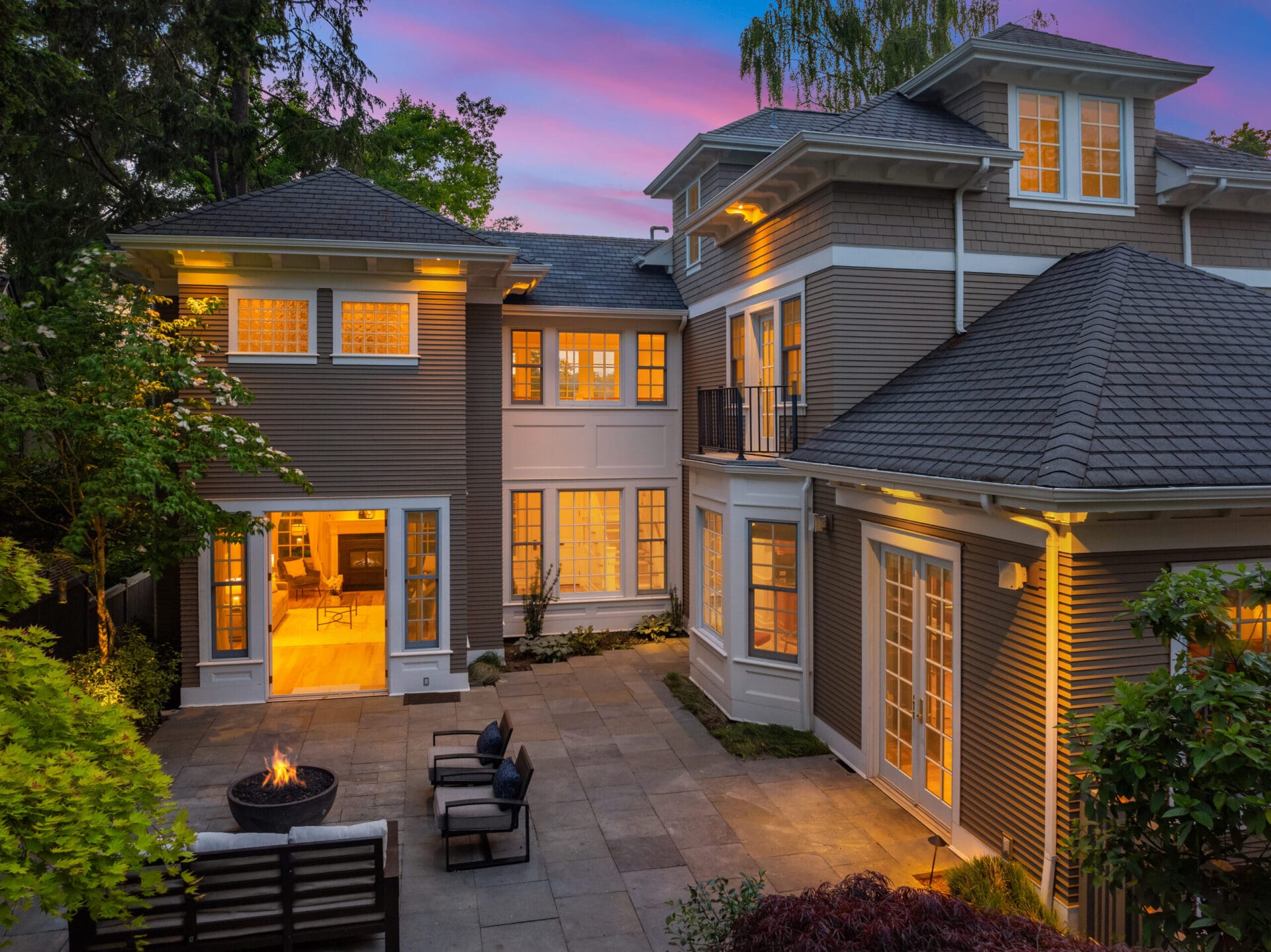 A large two-story house with several windows illuminated from within, showcasing a warm, inviting interior. The exterior features stylish shingles and off-white trim, and a well-maintained patio area with outdoor seating around a lit fire pit. The sky is vibrant at dusk.