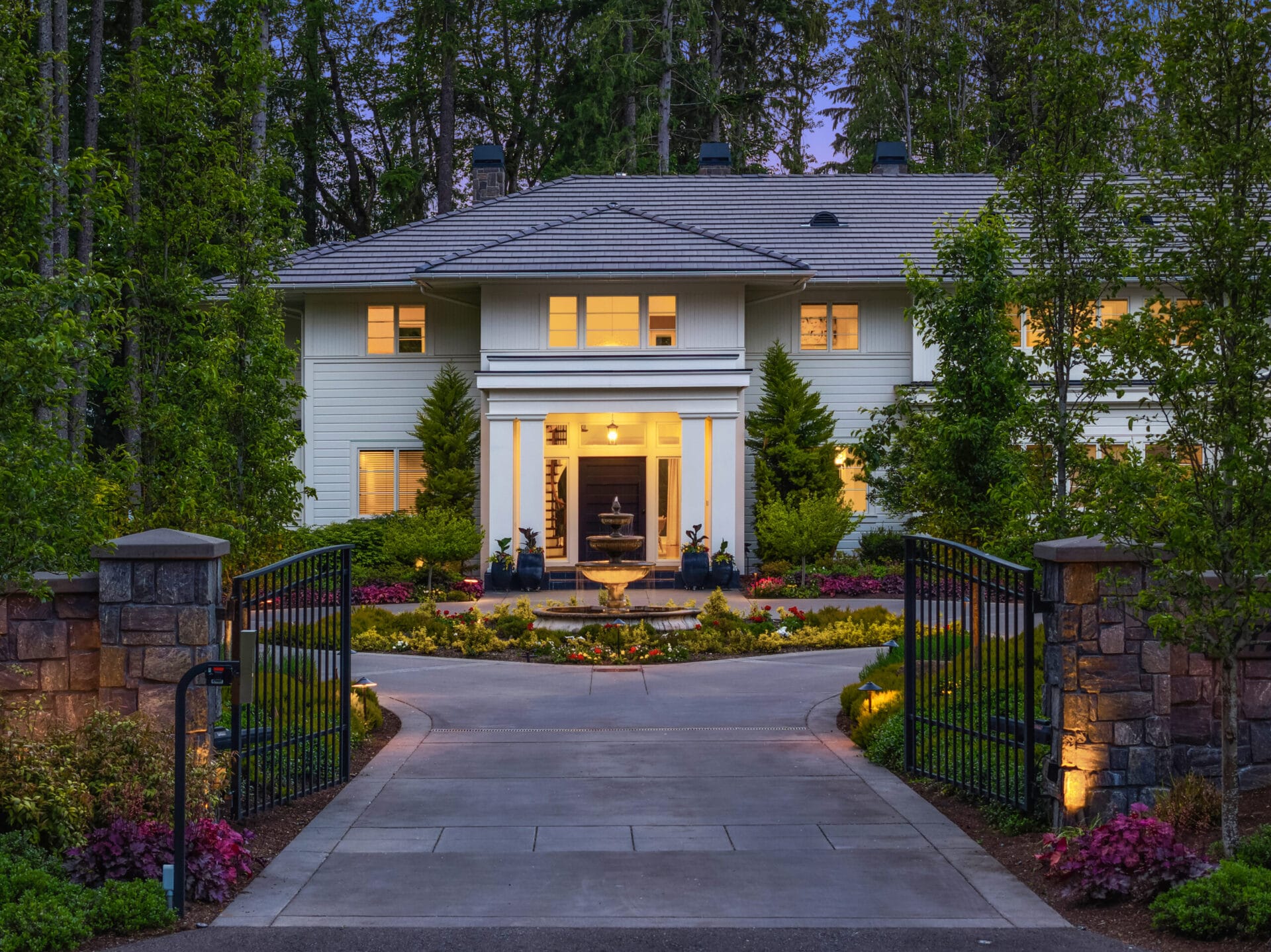 A large, two-story white house, illuminated at dusk, surrounded by lush greenery. The elegant entrance features tall windows, a central door, and a welcoming fountain. An open ornate gate and a neatly landscaped driveway lead to the front of the residence.