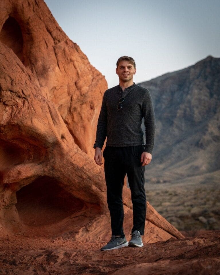 A person stands smiling on reddish-orange rock formations with a mountainous backdrop. They are wearing a dark long-sleeve shirt, black pants, and gray sneakers. The sky is clear. The setting appears to be a desert landscape at either sunrise or sunset.