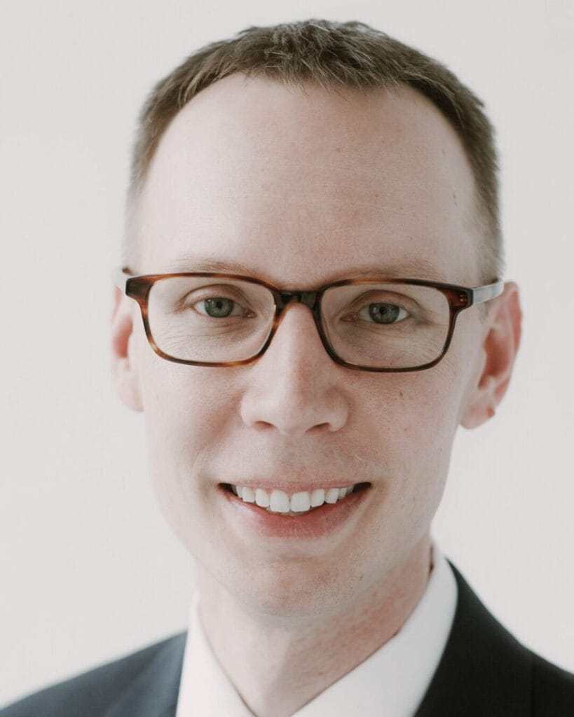 A man with short light brown hair, wearing glasses with dark frames, is smiling. He is dressed in a white shirt and a dark suit jacket, set against a plain light-colored background.