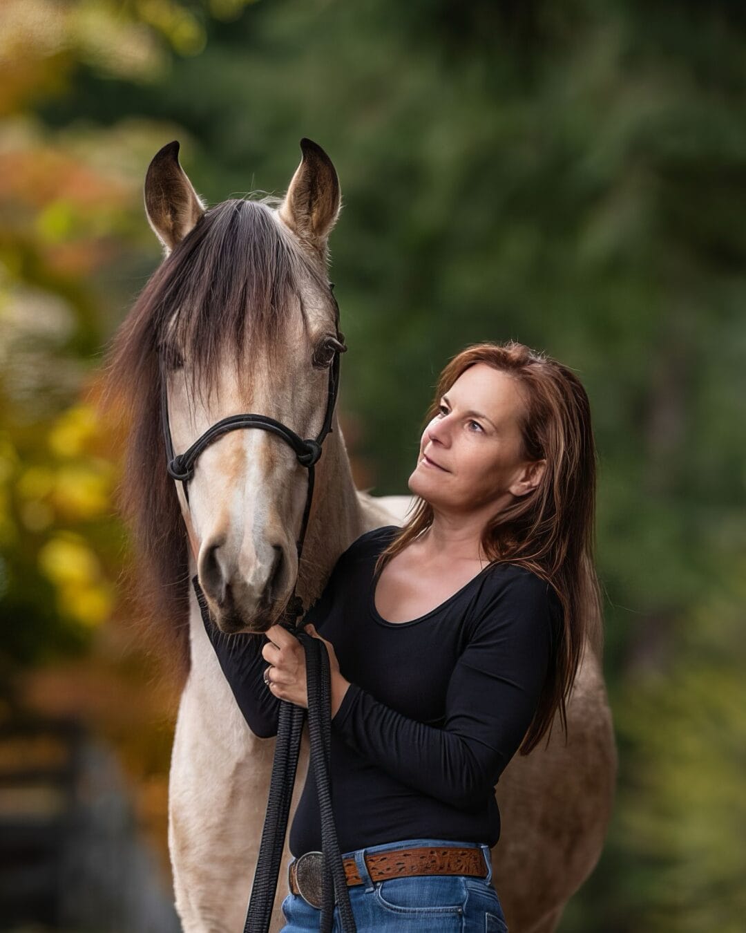 A woman with long, brown hair wearing a black top and jeans stands closely beside a light-colored horse. She holds the horse's reins and looks thoughtfully into the distance. The background is a lush, green, natural setting with soft, blurred foliage.