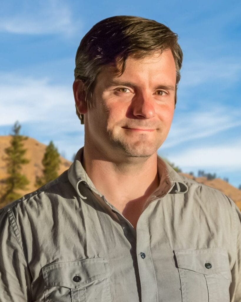 A man with short brown hair stands outdoors in front of a backdrop of trees and hills. He is wearing a light grey button-up shirt and is smiling slightly while looking at the camera. The sky behind him is blue with some wisps of clouds.