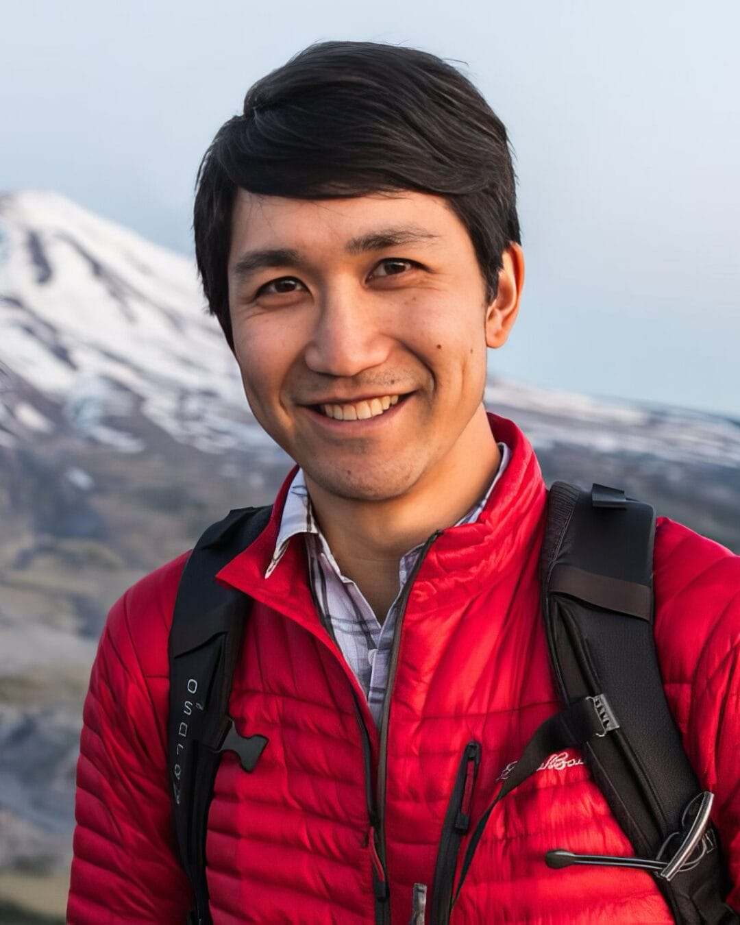 A person with dark hair smiles at the camera while standing outdoors. They are wearing a red jacket and have a black backpack. In the background, there is a snow-capped mountain and a blue sky.